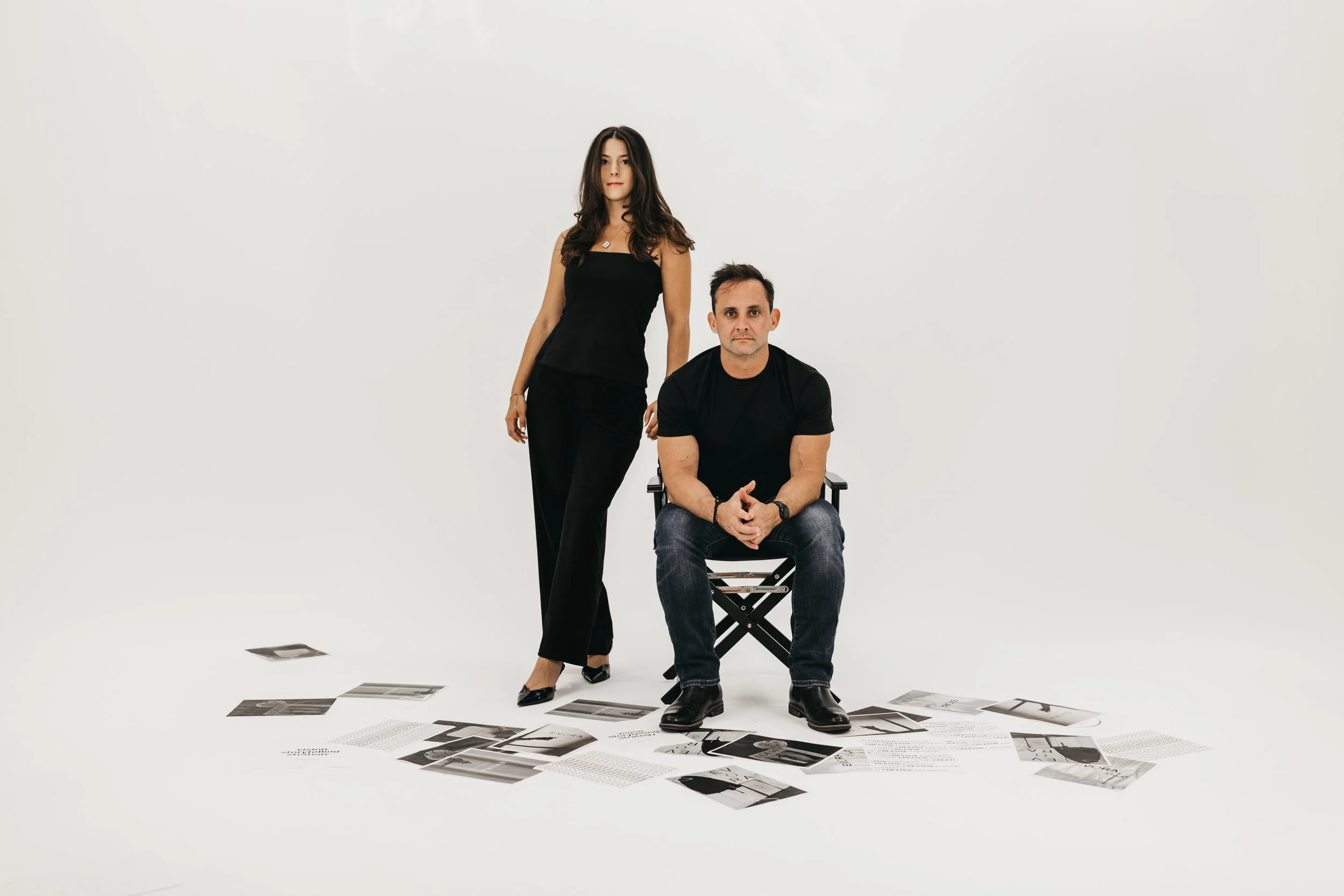 A woman standing next to a man sitting on a chair in a studio with white background, surrounded by scattered black-and-white photographs and papers on the floor.