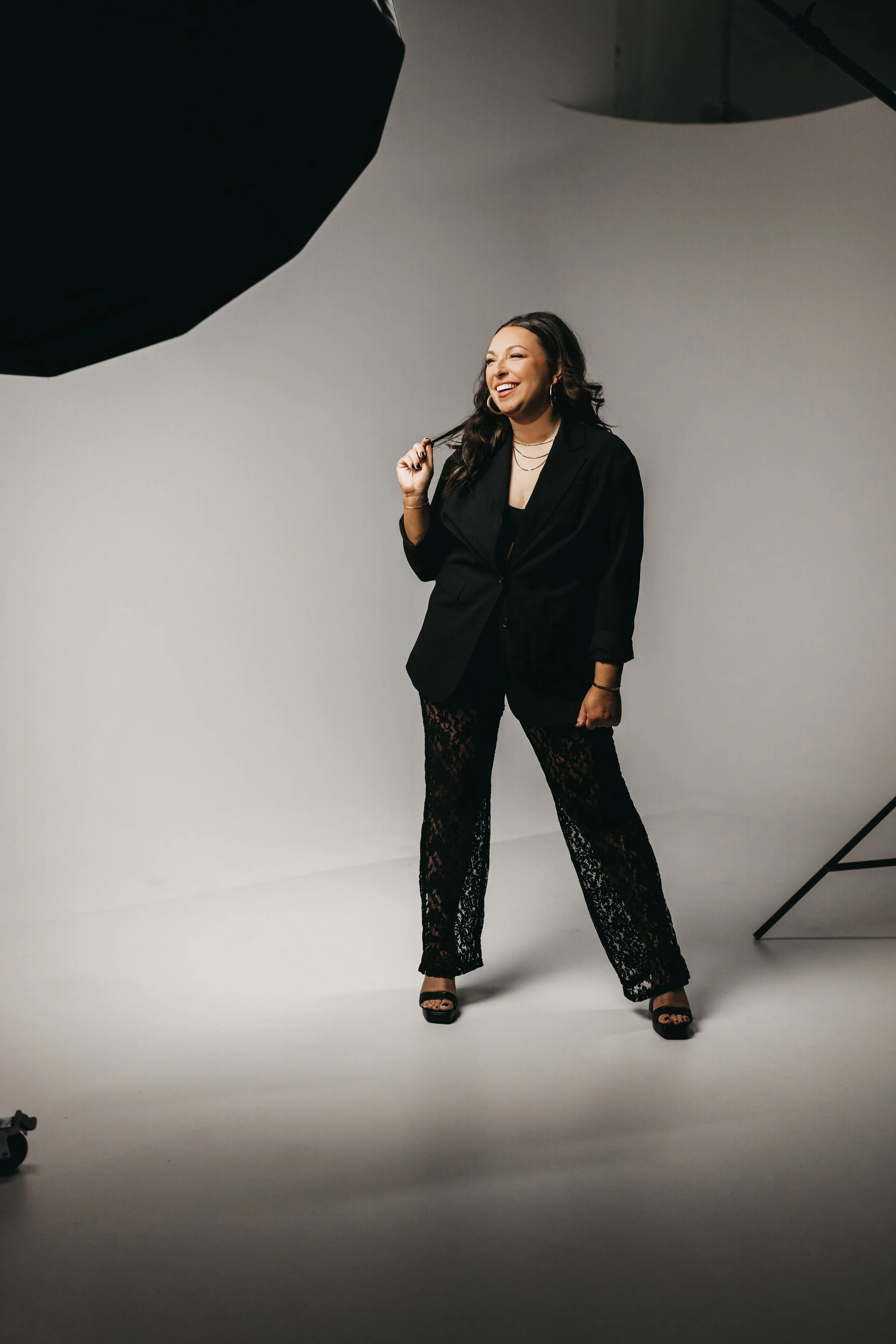 A woman smiling with long dark hair wearing a black blazer, lace pants, and high heels in a photography studio.