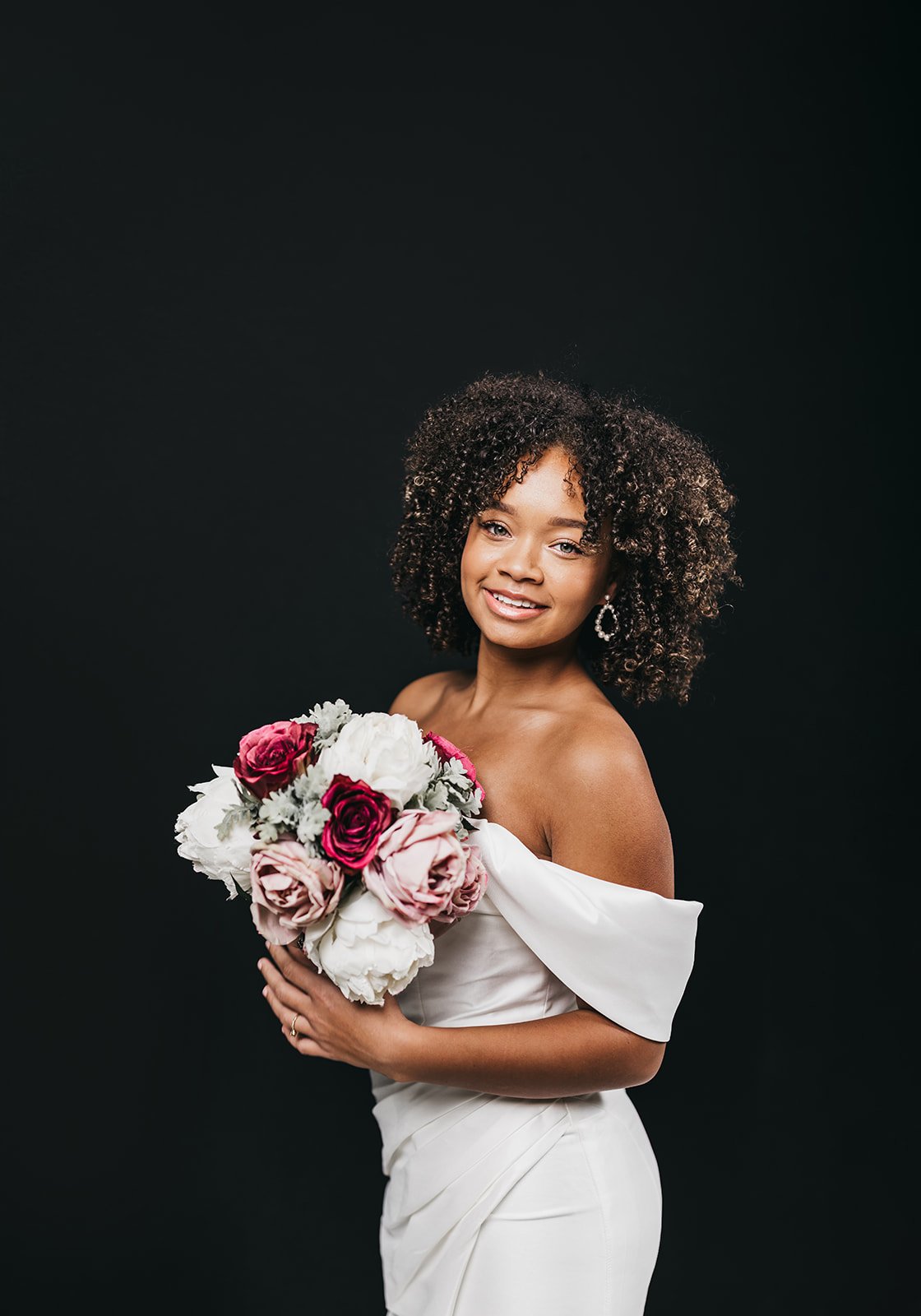 A woman in a white off-shoulder dress holding a bouquet of pink and white roses on a black background.