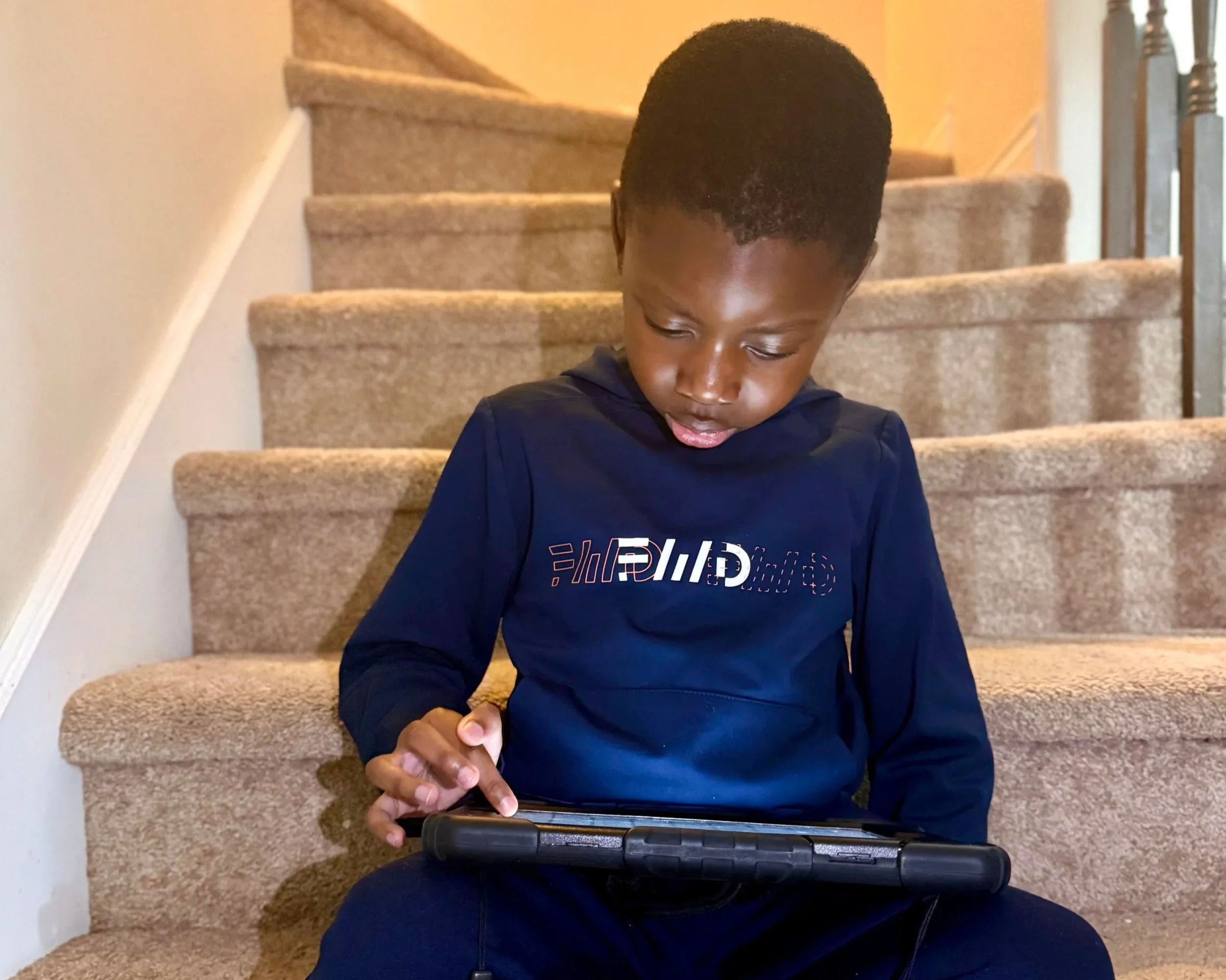 A child named CJ sitting on stairs in his home, using an Ipad