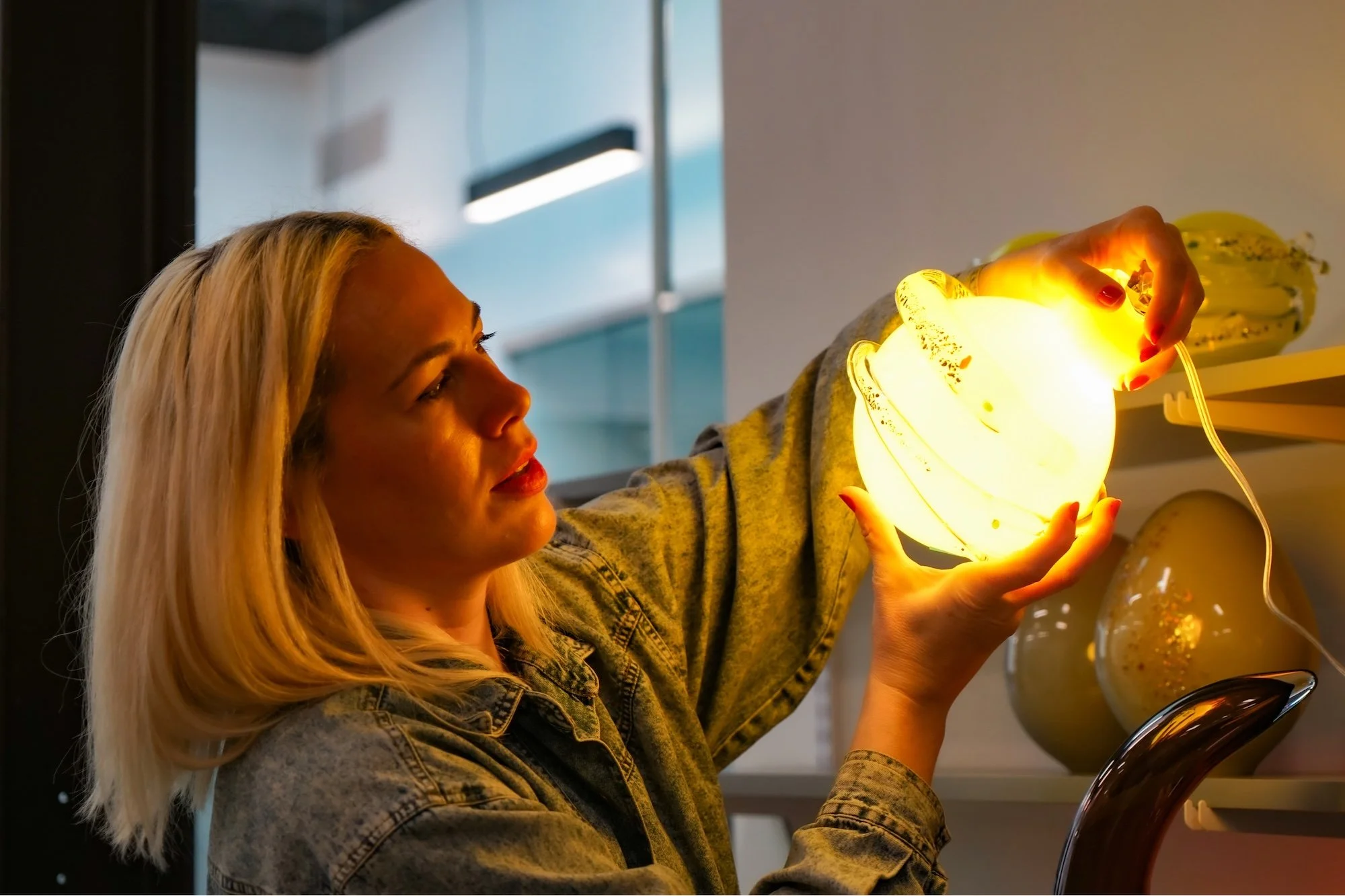 A young woman with blonde hair holding a glowing yellow decorative lamp in a store.