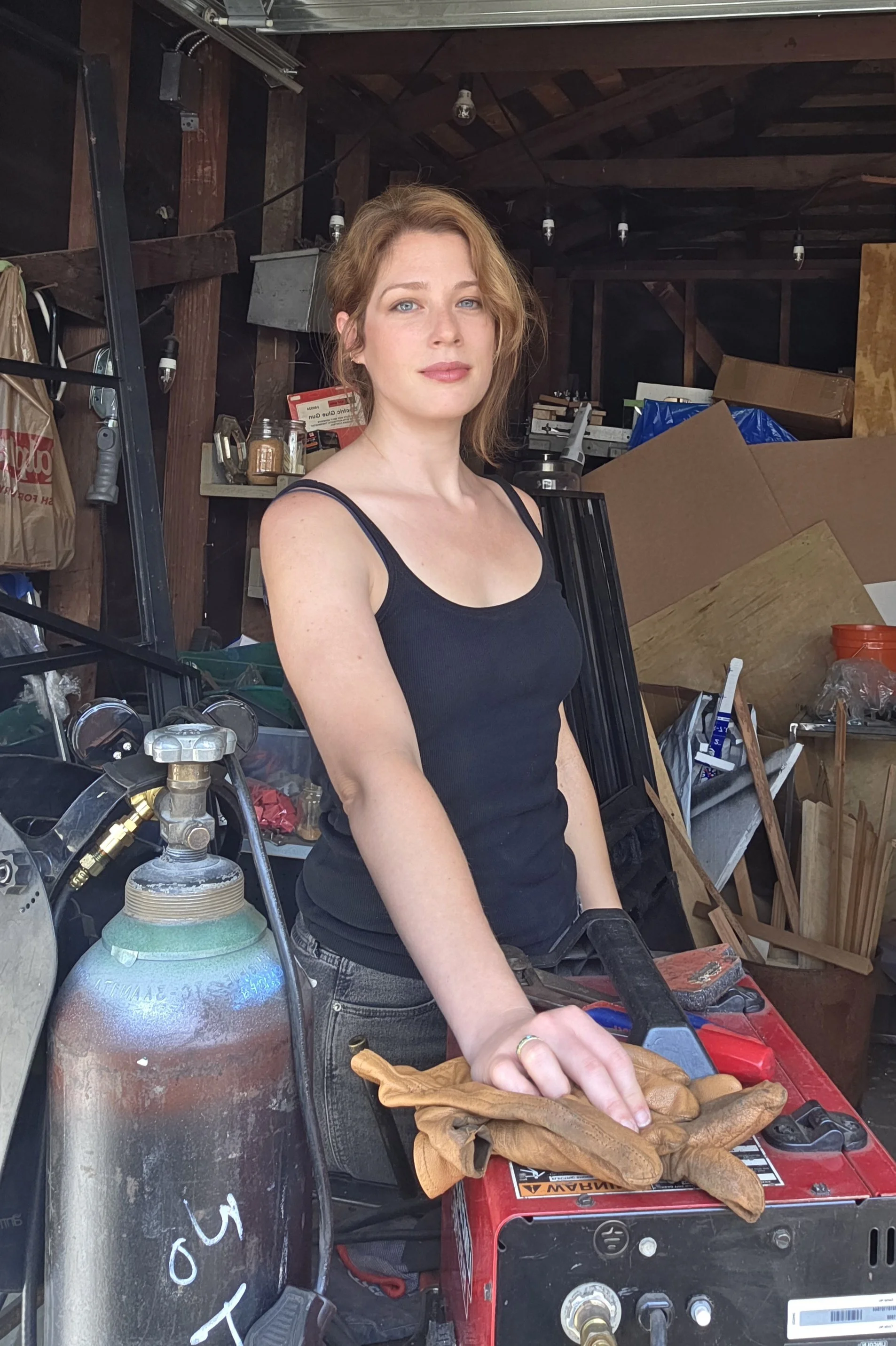 A woman with red hair wearing a black tank top standing in a garage surrounded by tools, equipment, and a large propane tank, with a welding machine in front of her.