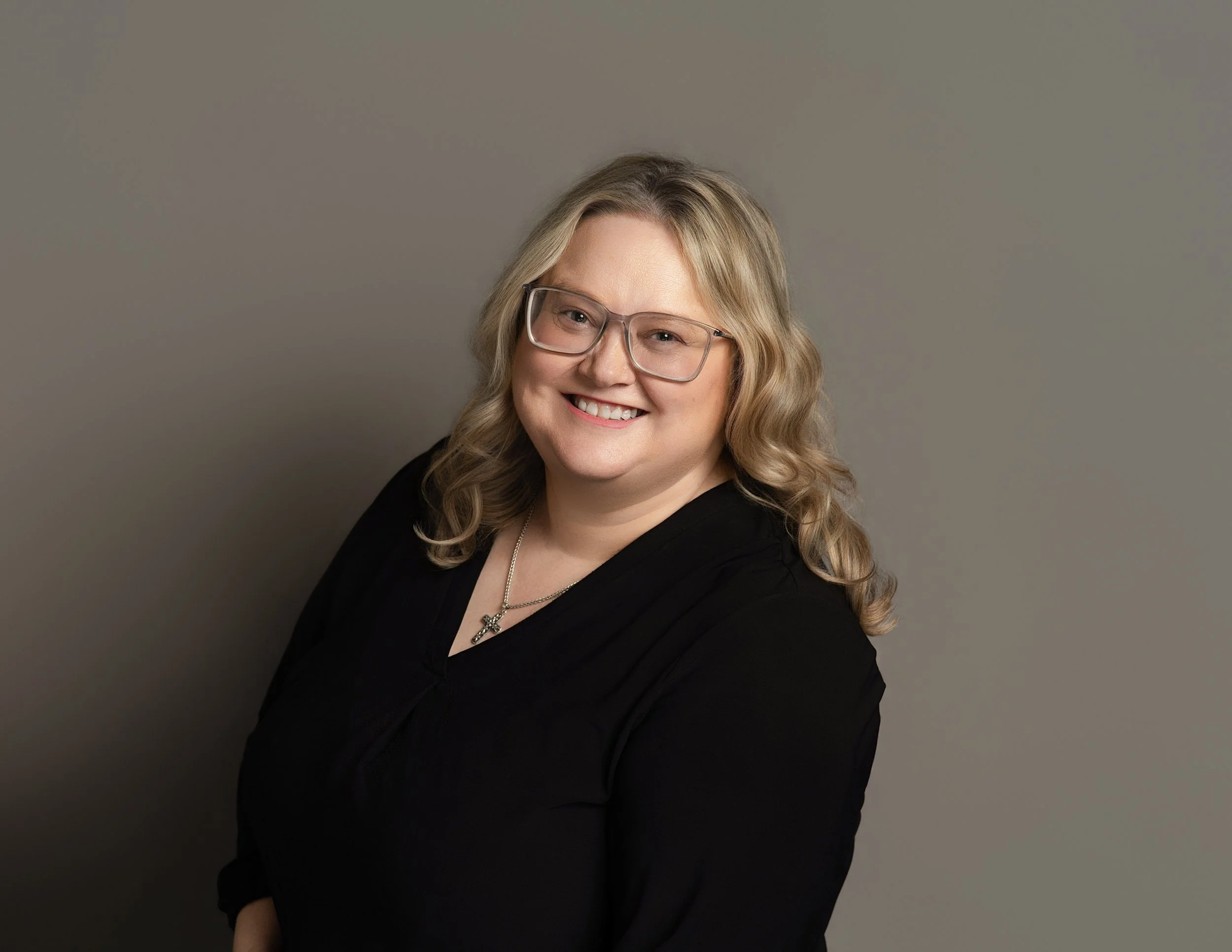 A woman with wavy blonde hair and glasses smiling at the camera, wearing a black outfit and a star-shaped necklace against a plain grey background.