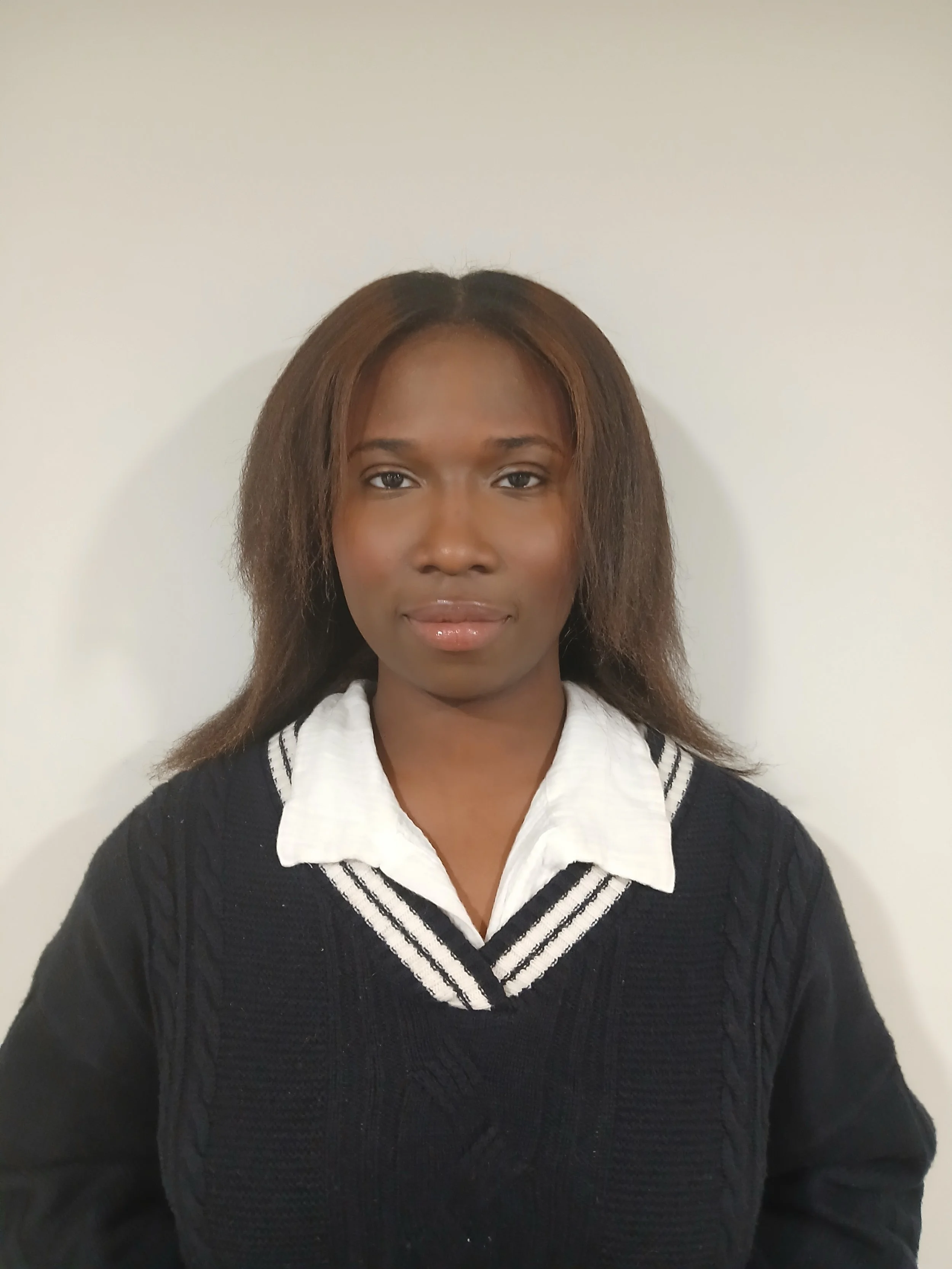 A woman with straight brown hair wearing a white collared shirt and a black sweater with white stripes near the collar, standing against a plain light-colored wall.