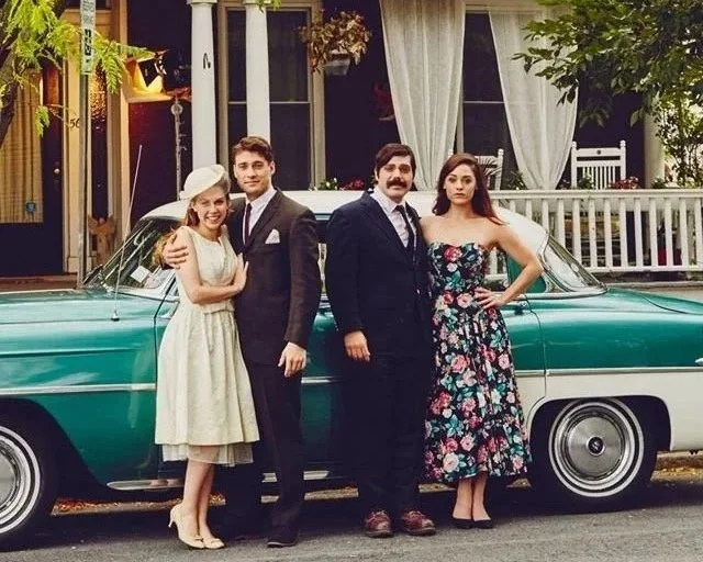Four people standing in front of a vintage teal car, two men and two women, at an outdoor setting with a house and porch in the background. The woman on the left is wearing a light-colored dress and hat, the man beside her is in a dark suit, the man