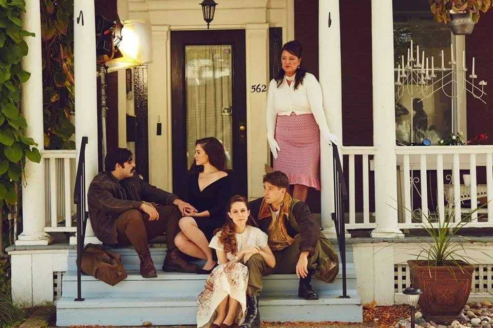 Five young people sitting and standing on the front porch of the scariest house in America, with two women and three men, engaged in conversation, with decorative plants and a tree nearby.