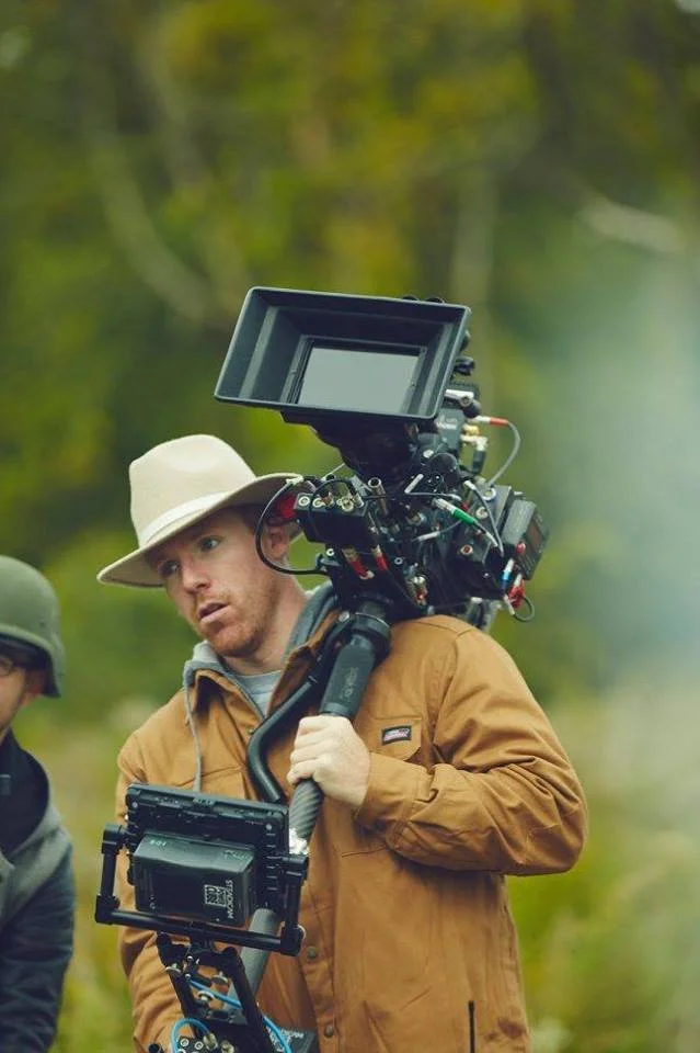 Filmmaker on an Upstate NY film location operating a camera on a shoulder rig outdoors with trees in the background.