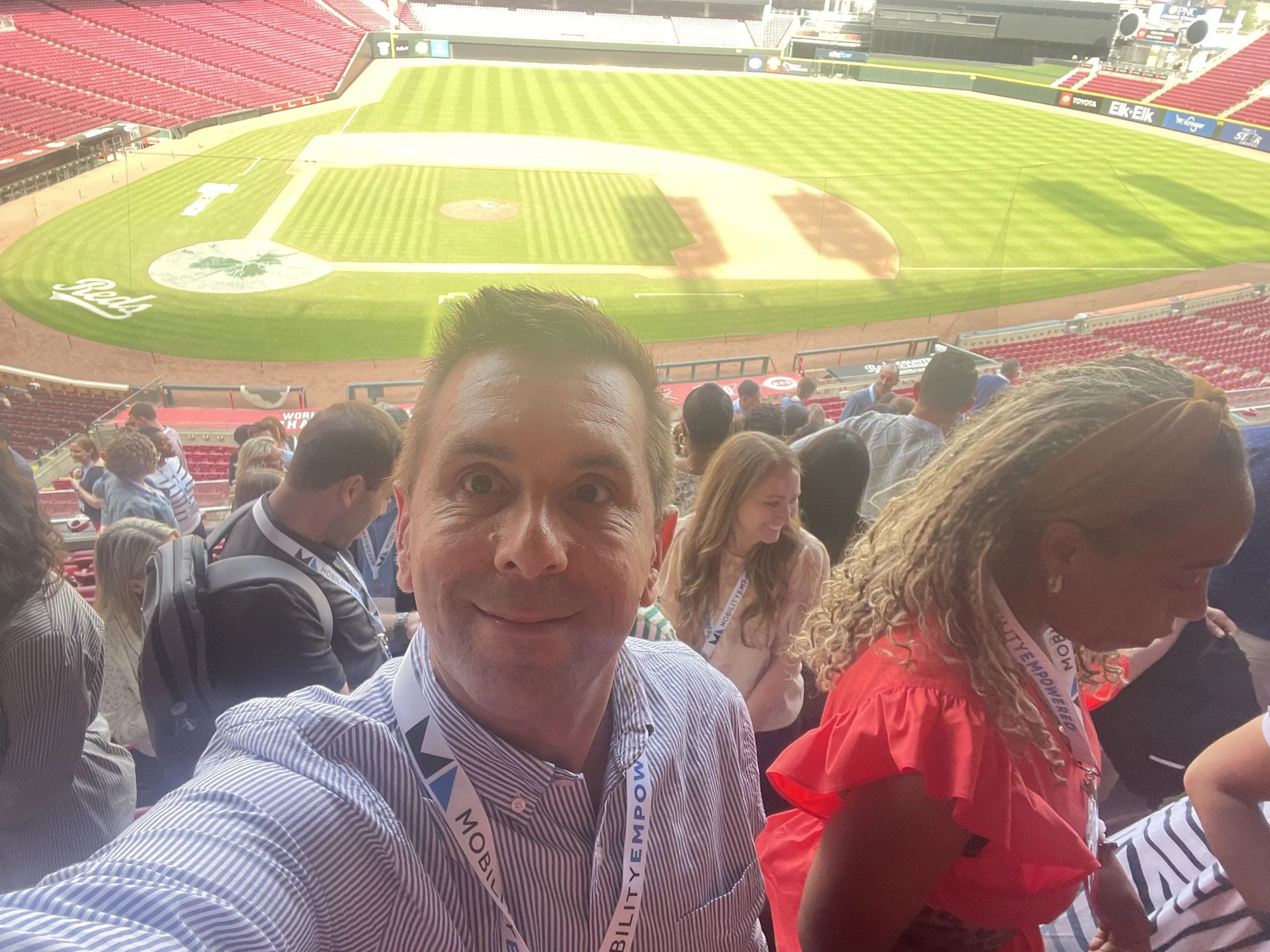 People sitting in stadium seats overlooking a baseball field, with a man taking a selfie in the foreground.