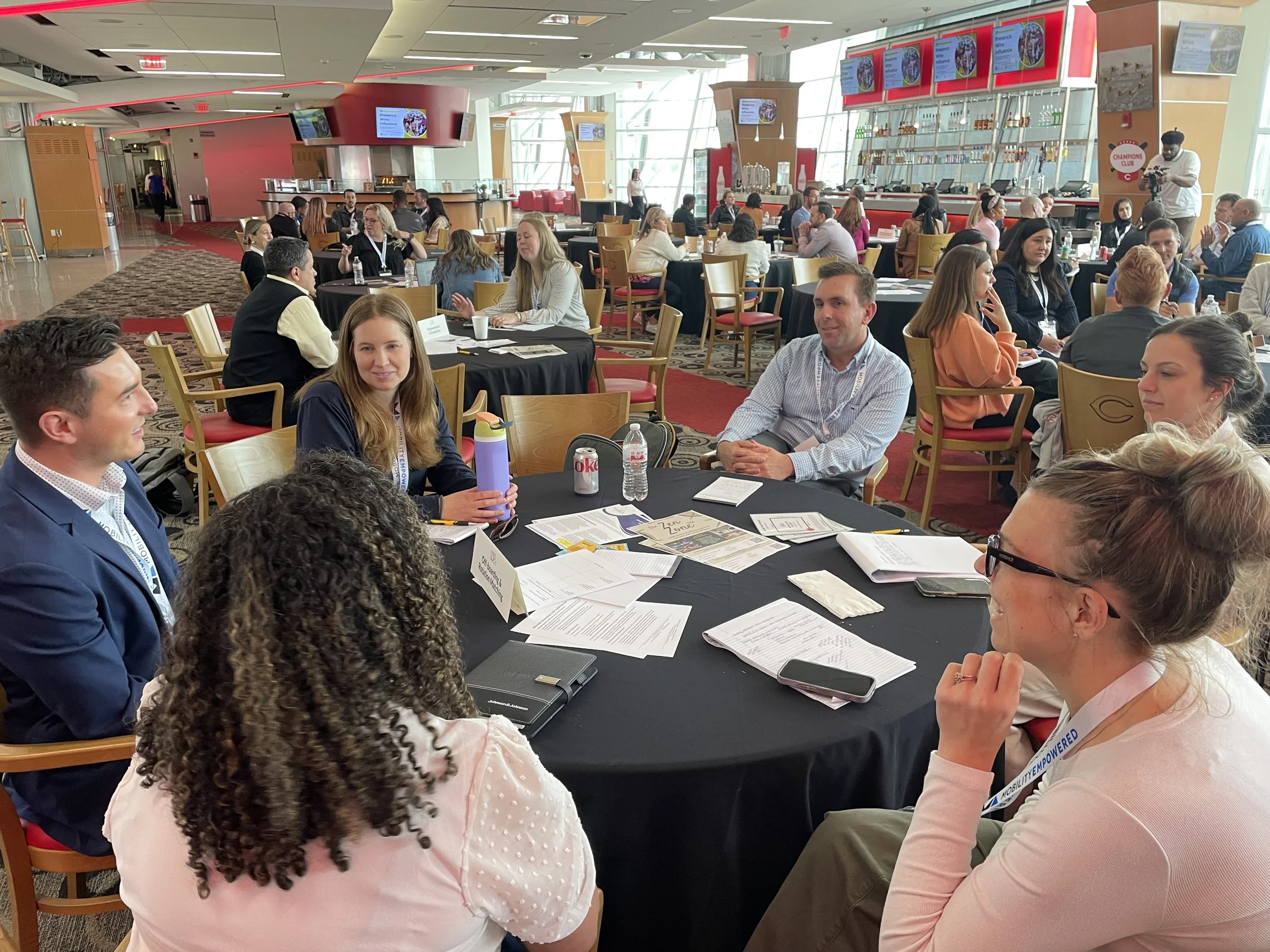 A group of diverse people sitting around a round table at a conference, engaging in a discussion, with a busy hospitality area in the background of a large, modern convention center.