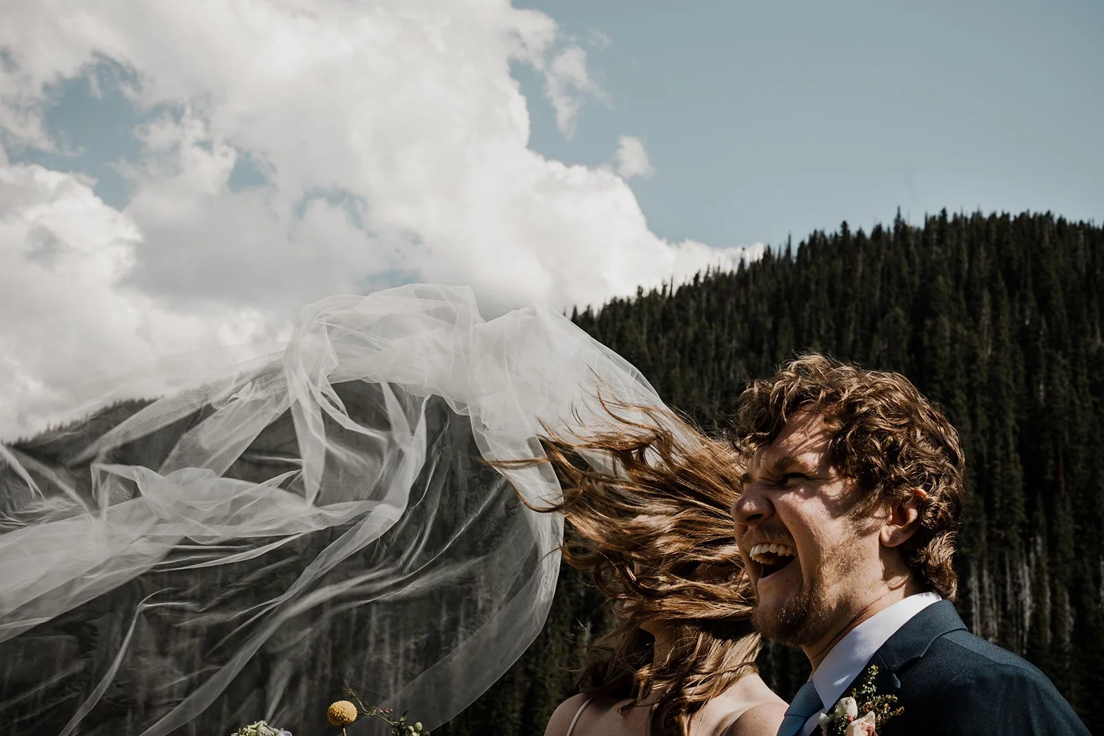 A bride's veil blows in the wind while the groom laughs at their Manning Park mountain wedding. Wedding photography by Sea to Sky Studios.