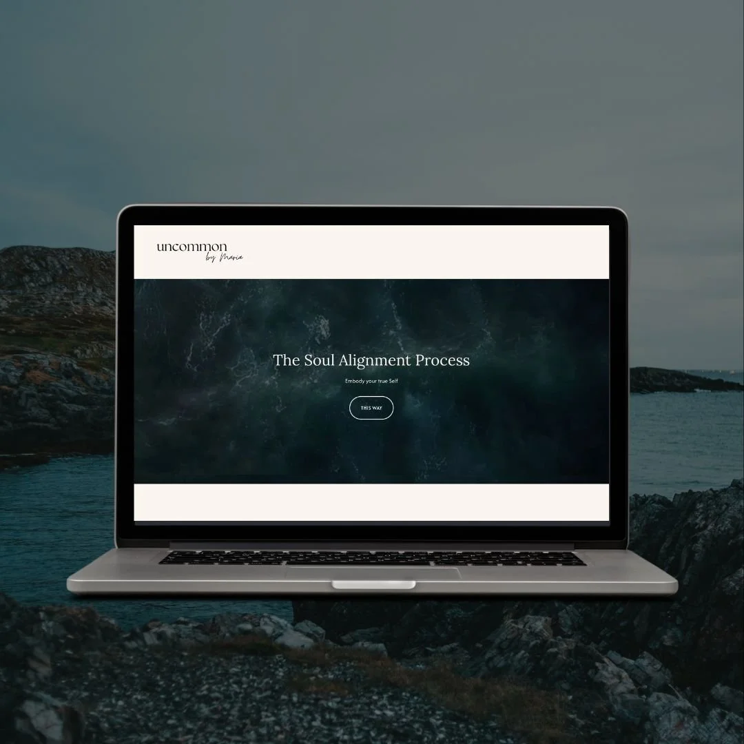 Laptop displaying a webpage titled 'The Soul Alignment Process' by Uncommon on a rocky shoreline with water and mountains in the background.