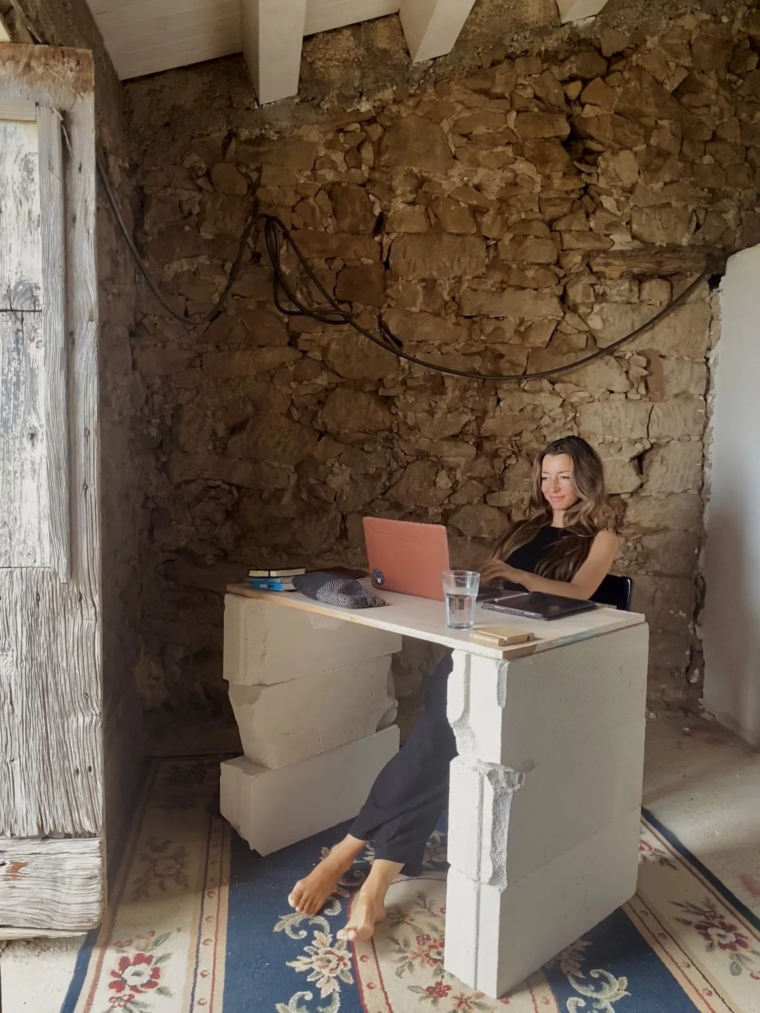 A woman with long hair working on a laptop at a makeshift desk made of large stone blocks, inside a rustic room with exposed stone wall, wooden beam ceiling, and a patterned rug on the floor.