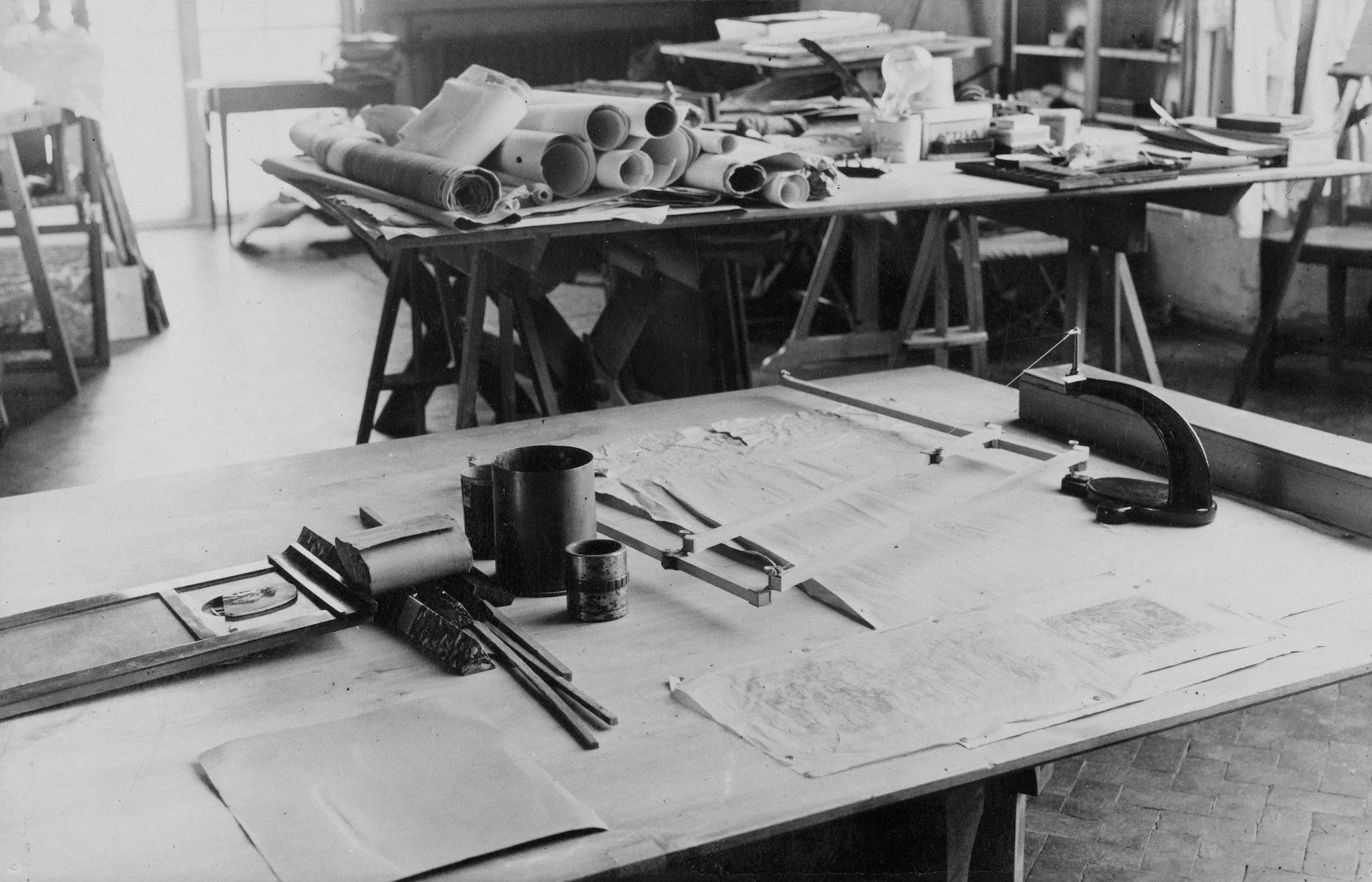 A workshop with tables holding architectural drawings, rulers, and tools, and shelves in the background with rolled-up blueprints and papers.