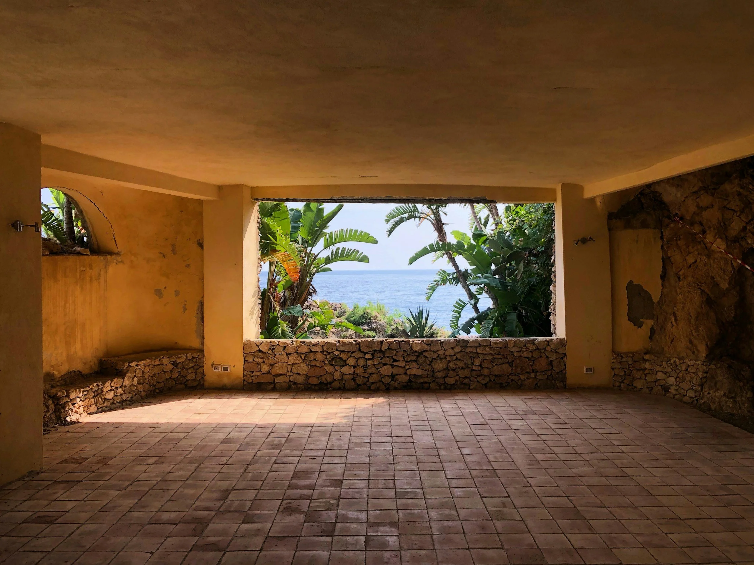 A covered patio or terrace with a stone wall and large open window, overlooking a lush tropical garden with large green leaves, palm trees, and the ocean in the background.