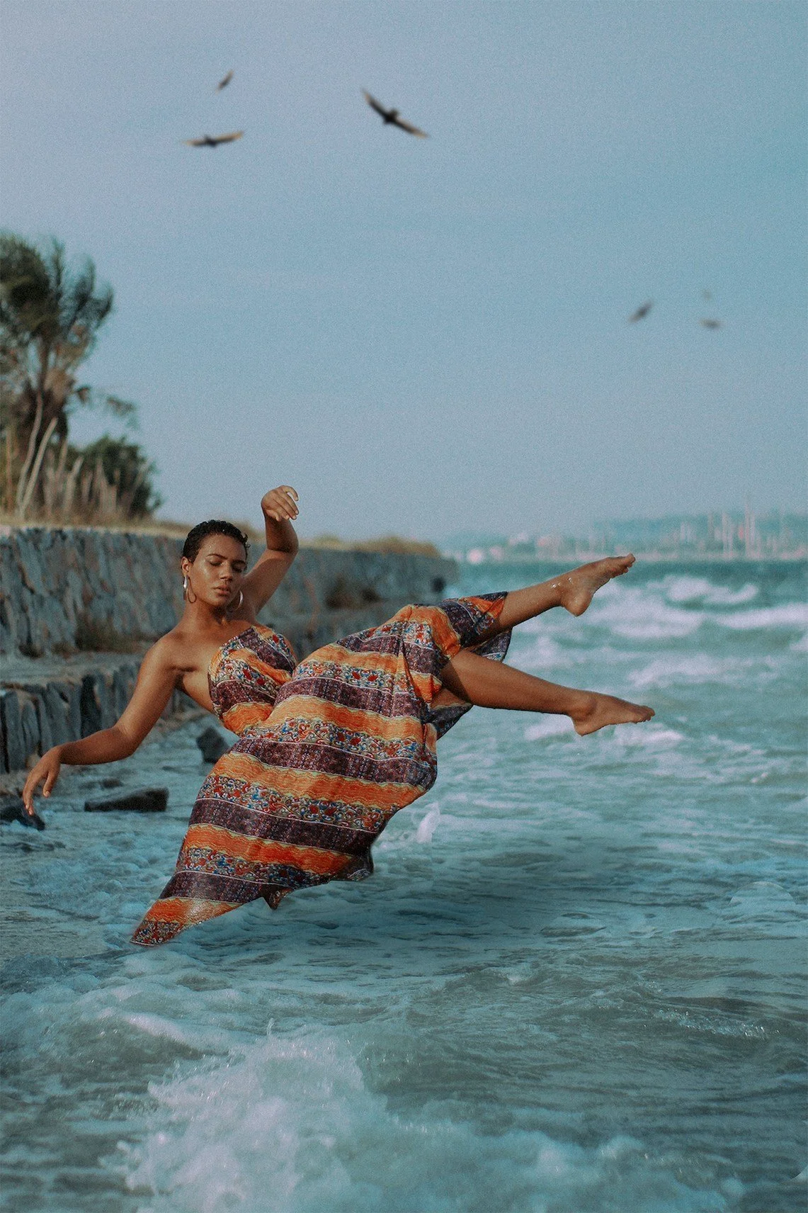 Woman in a colorful patterned dress kicking toe in the ocean water near a rocky shoreline with birds flying overhead.