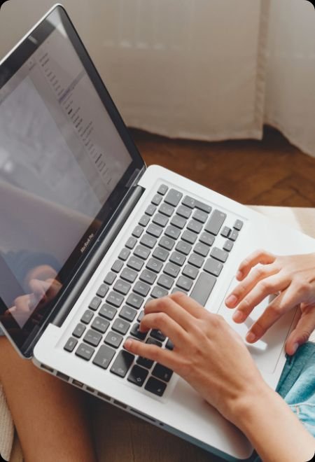 Person typing on a silver laptop with a black keyboard, on a white surface, in a room with a wooden floor and light-colored curtains.