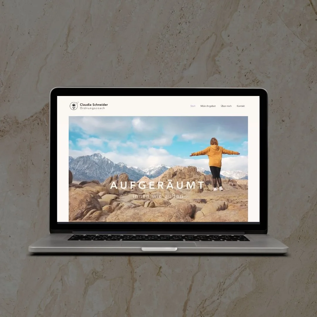 A laptop on a marble surface displaying a website with a woman standing on rocks in a mountain landscape with arms outstretched, wearing a yellow jacket. The website has German text and a menu at the top.
