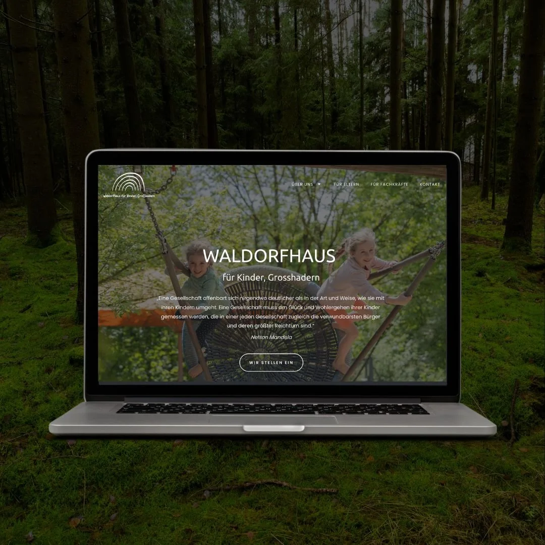 A laptop displaying a website about a forest house for children and seniors, placed on the ground in a forest setting with trees and green moss.