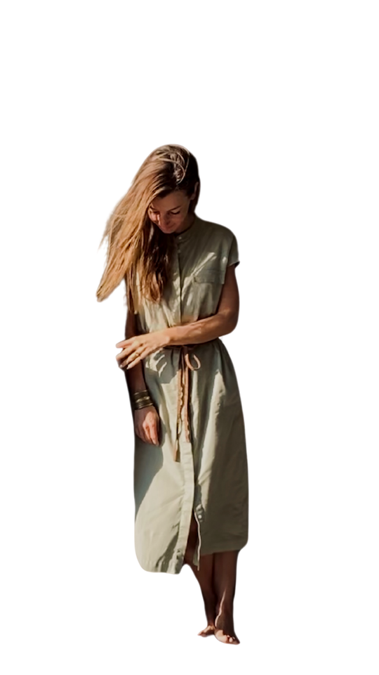 A woman with long blond hair wearing a light-colored, sleeveless, buttoned dress tied at the waist, barefoot, standing on a beach.