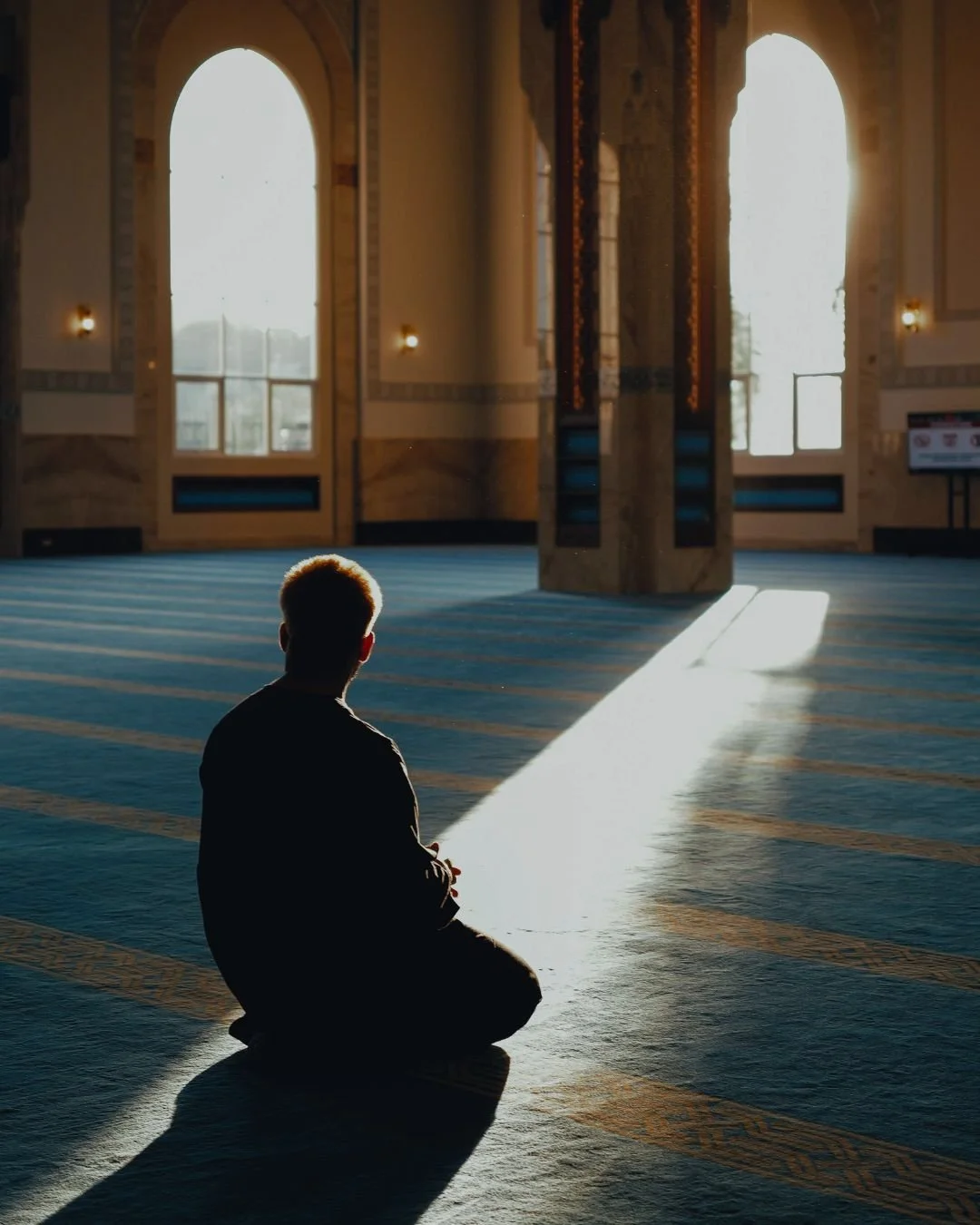 A person sitting in prayer or meditation on a carpeted floor of a prayer room or mosque, illuminated by sunlight streaming through large arched windows behind them.