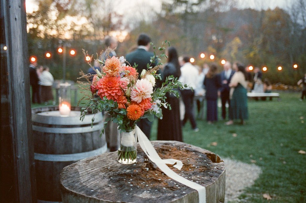 Wedding bouquet on barrel durinhg cocktail hour, with guests mingling candidly in background at tops'l farm in Maine