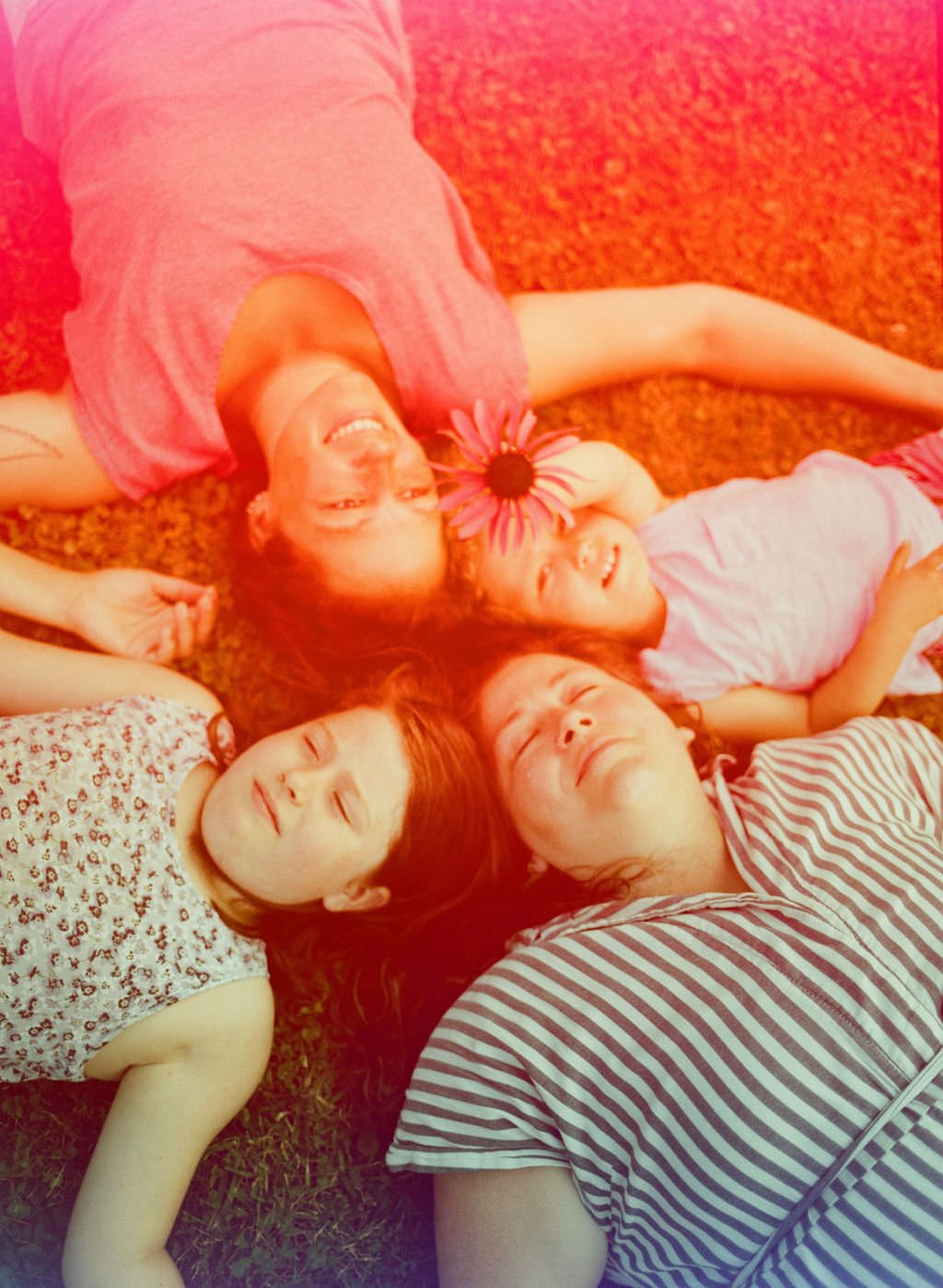 A queer family of 4 puts their heads together in their backyard on the grass smiling looking up, theres a colorful light leak across the frame making the image pink and rainbows