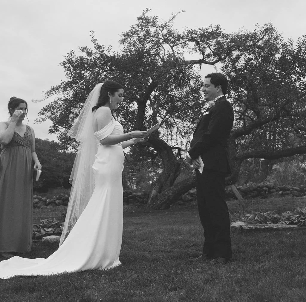 Black and white film photography of couple exchanging vows with sister wiping tear in background under trees of tops'l farm in Maine