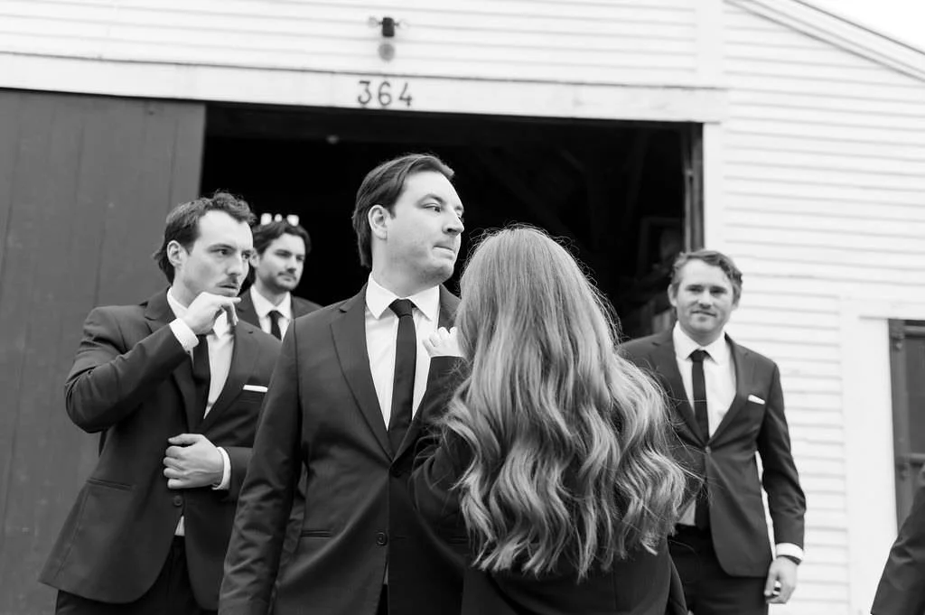 The groom looks off while someone fixes his pocket square, with his friends in the background, on black and white film