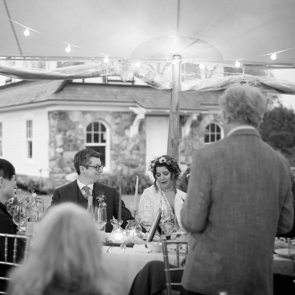 the father of the groom gives a speech under a tent in southern maine wedding during this tender moment where the bride and groom look at eachother during dinner