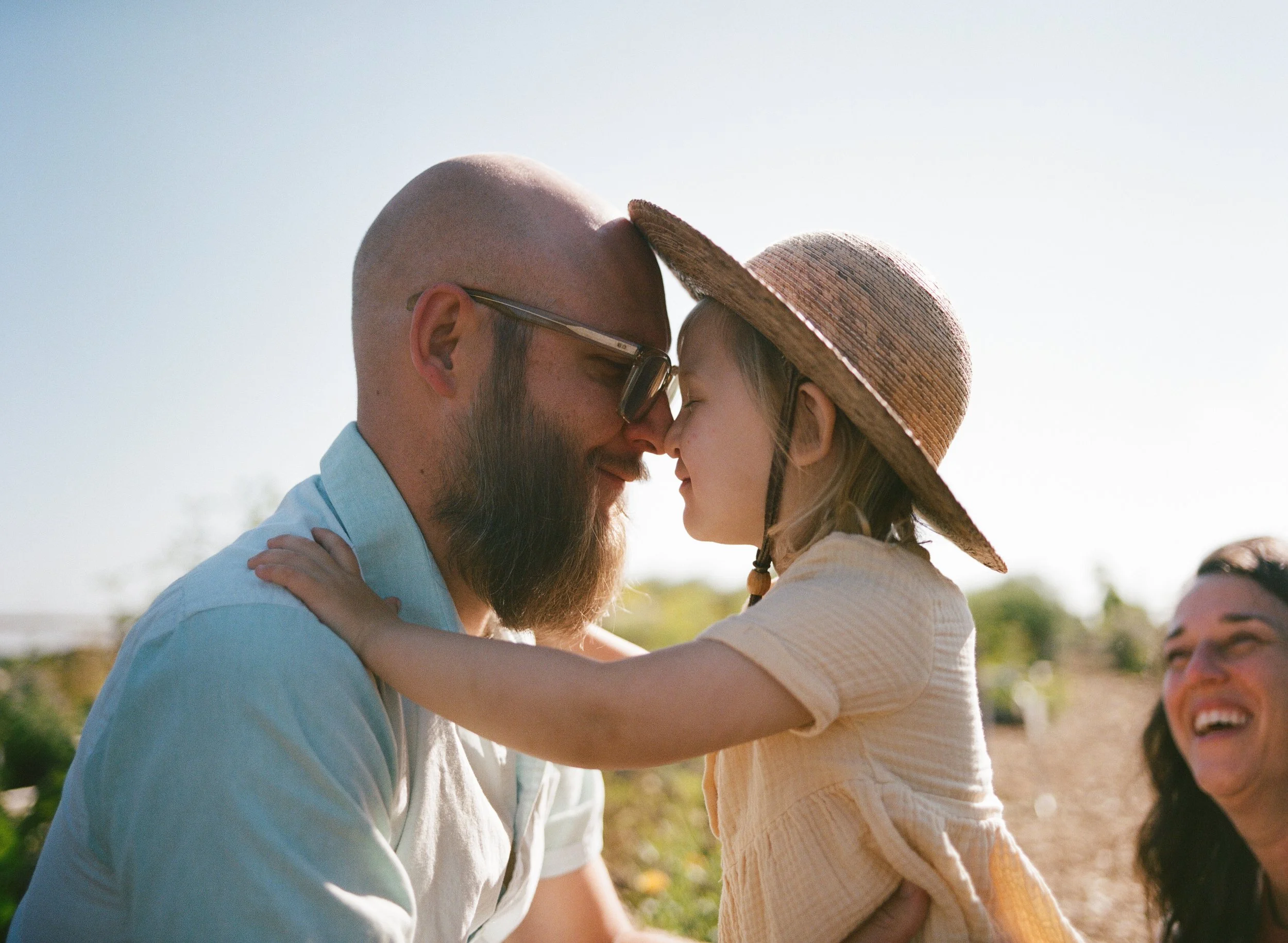 A dad and daughter touch noses at their local community garden in portland maine while mom looks on smiling during this candid documentary photo session