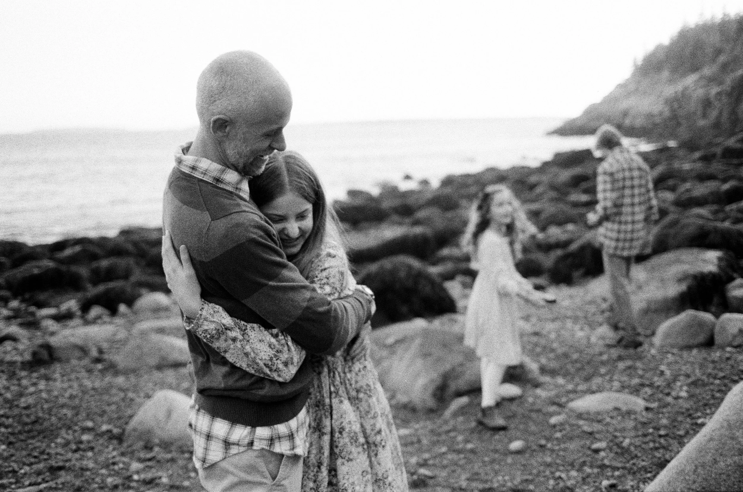 A daughter and dad hug while the other kids play behind them at a rocky beach in Acadia