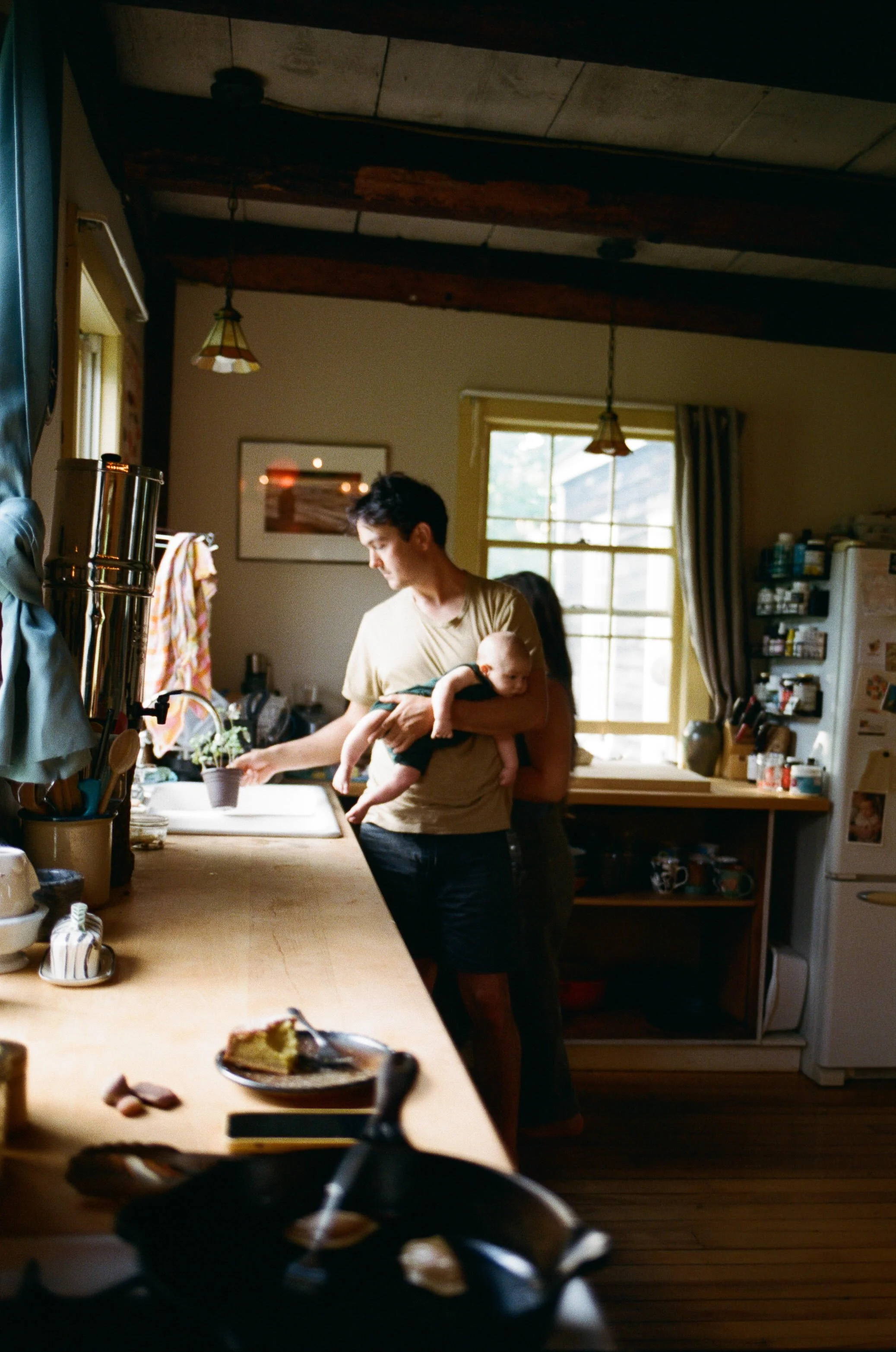 documentary family photography midcoast maine - a man water his plants in the kitchen while holding his newborn