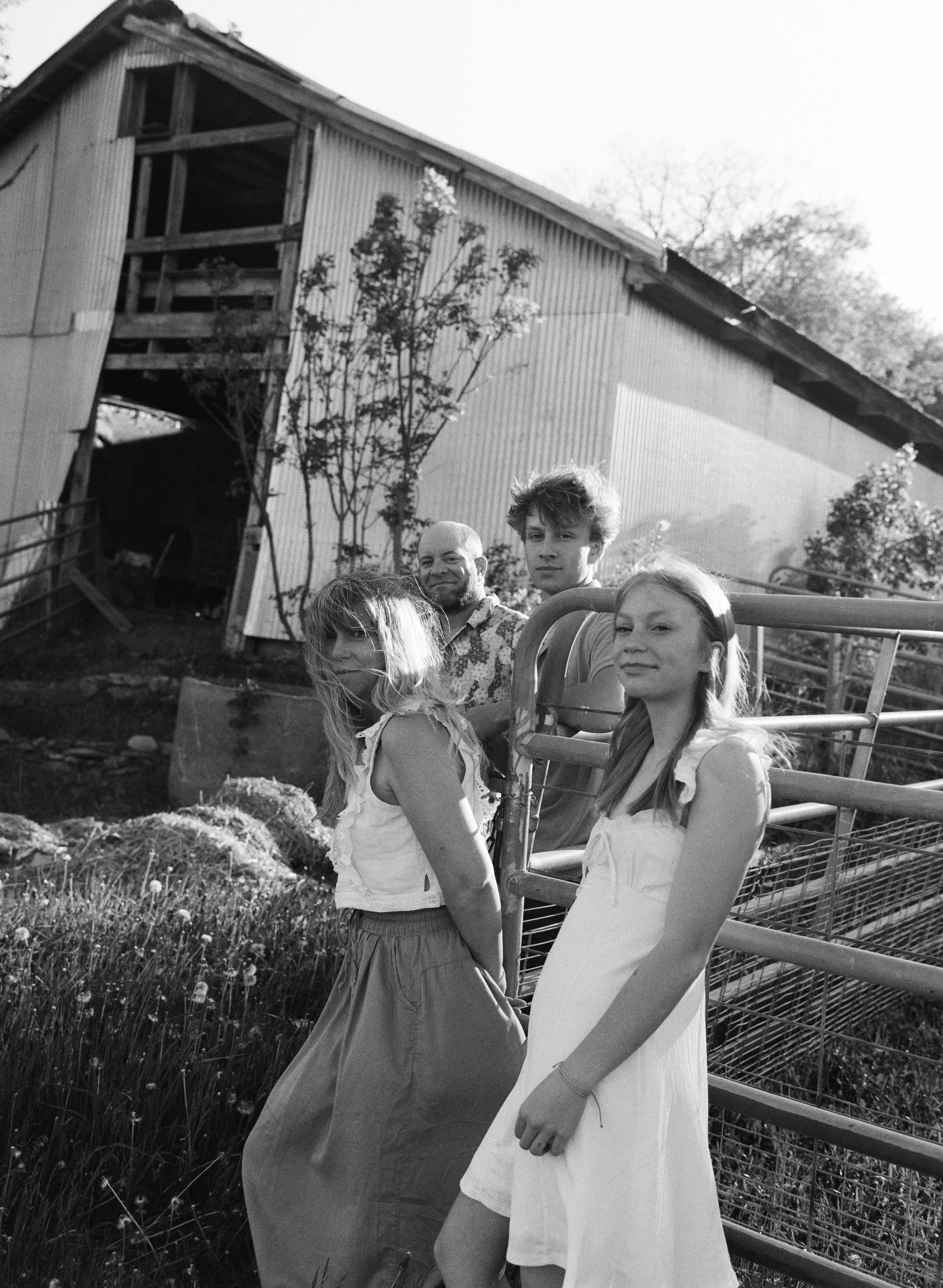 A family of 4 layered on a fence outside their barn at home in maine looking at the camera