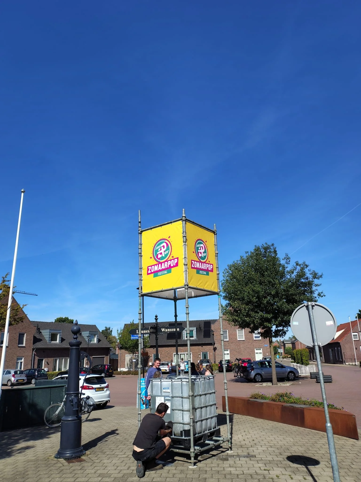 Mensen die werken aan een opbouw voor het Zomaarpop festival, met een grote gele banner en een blauwe lucht op een zonnige dag.