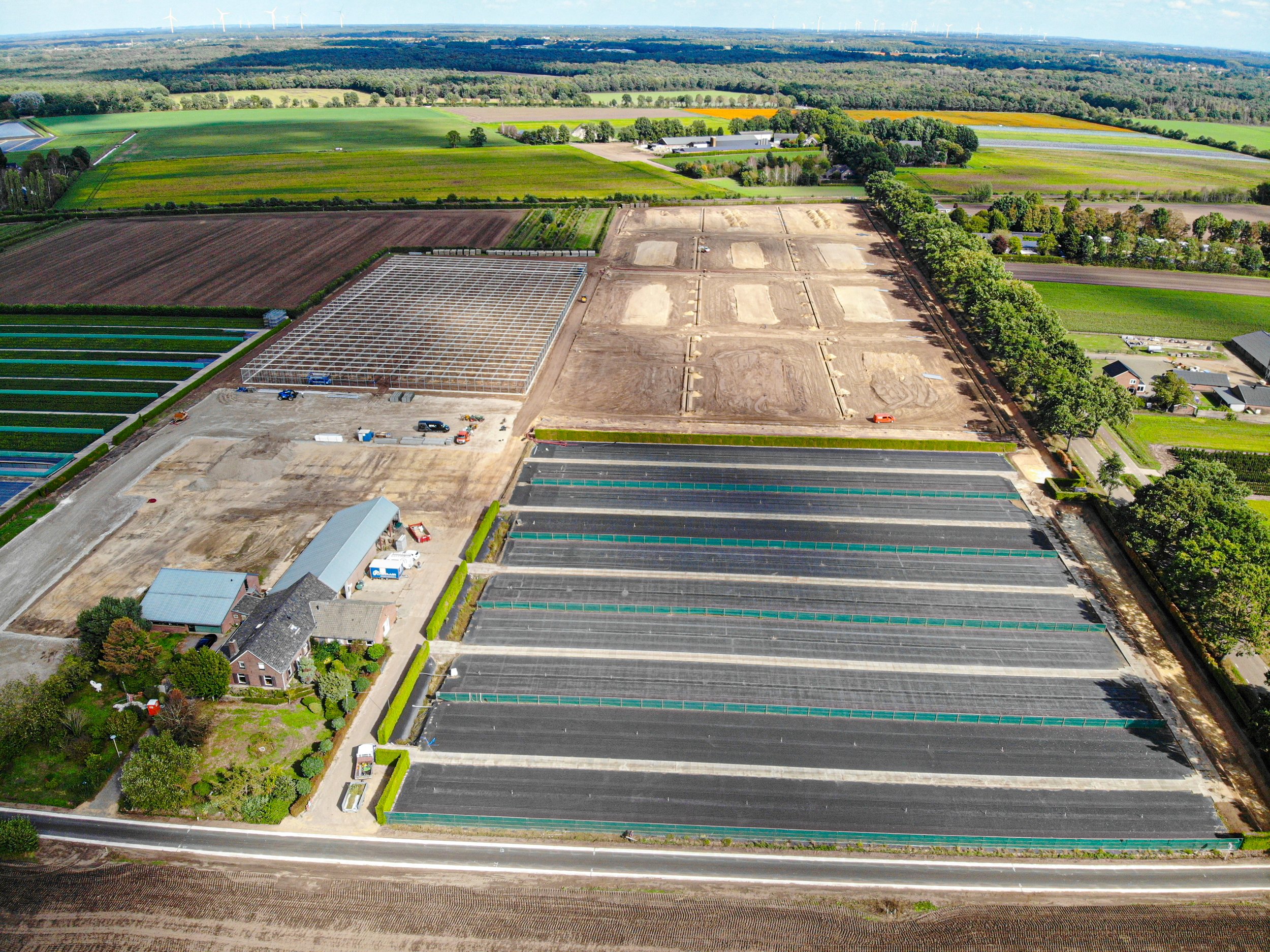 Luchtfoto van een landbouwgebied met verschillende gewassen, een kas, en windmolens op de achtergrond.