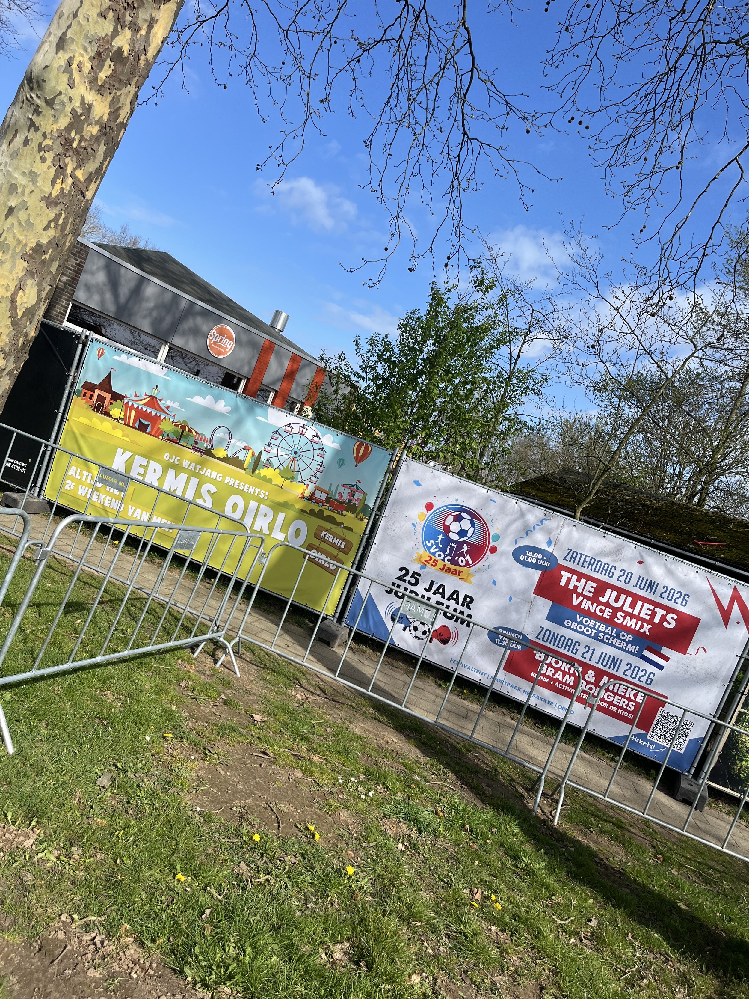 Een reclamebord voor een kermis en een sportevenement, met bomen en een blauwe hemel erboven.