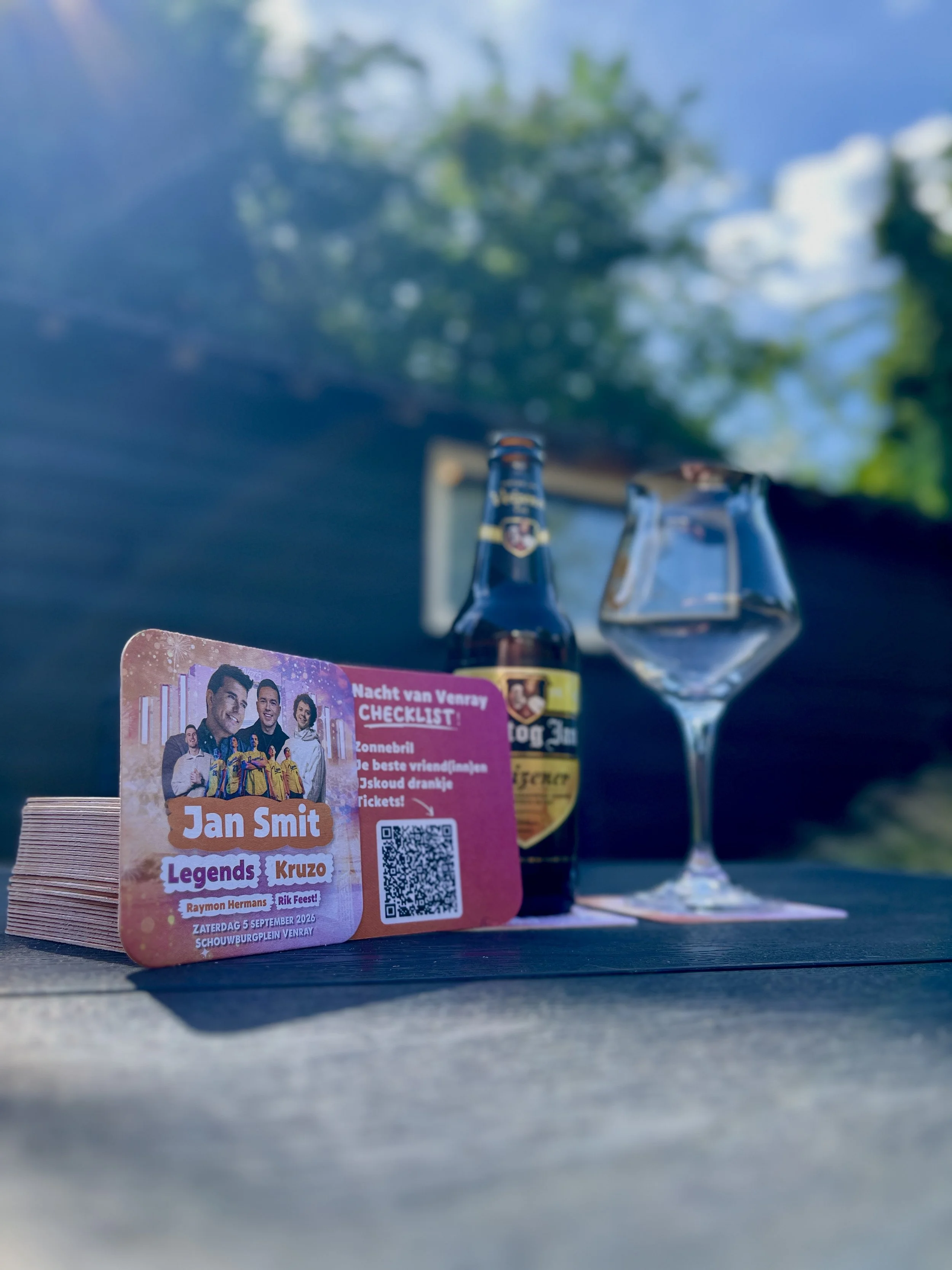 Evenementkaart en een bierflesje en een leeg wijnglas op een tafel, met een boom en een blauw hemel op de achtergrond.