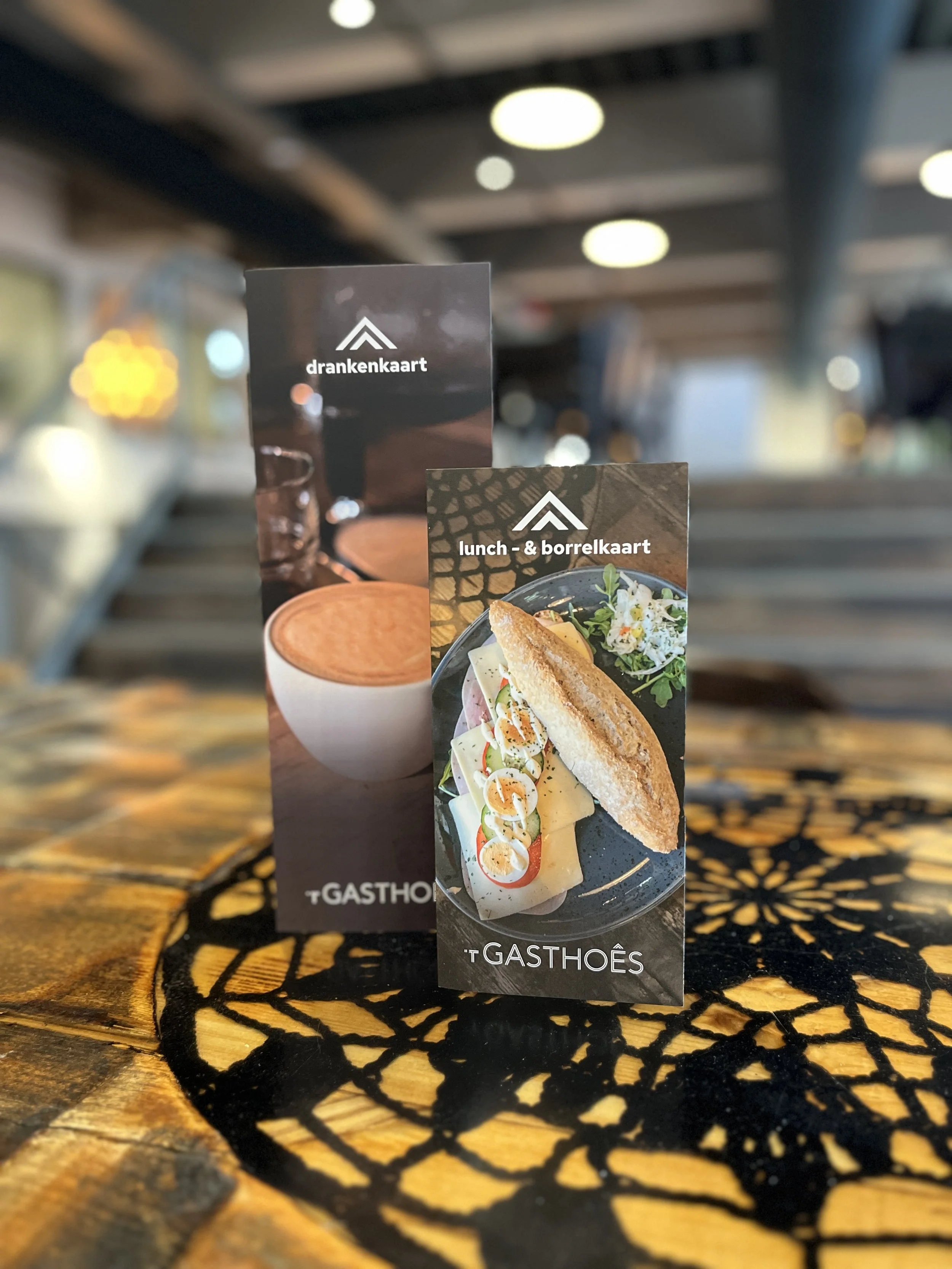 Een menu van een restaurant met de teksten 'drankenkaart' en 'lunch- & borrelkaart' en een afbeelding van een sandwich met salade, ei en tomaat op de voorgrond, geplaatst op een tafel met een zwart uitgesneden tafelkleed.