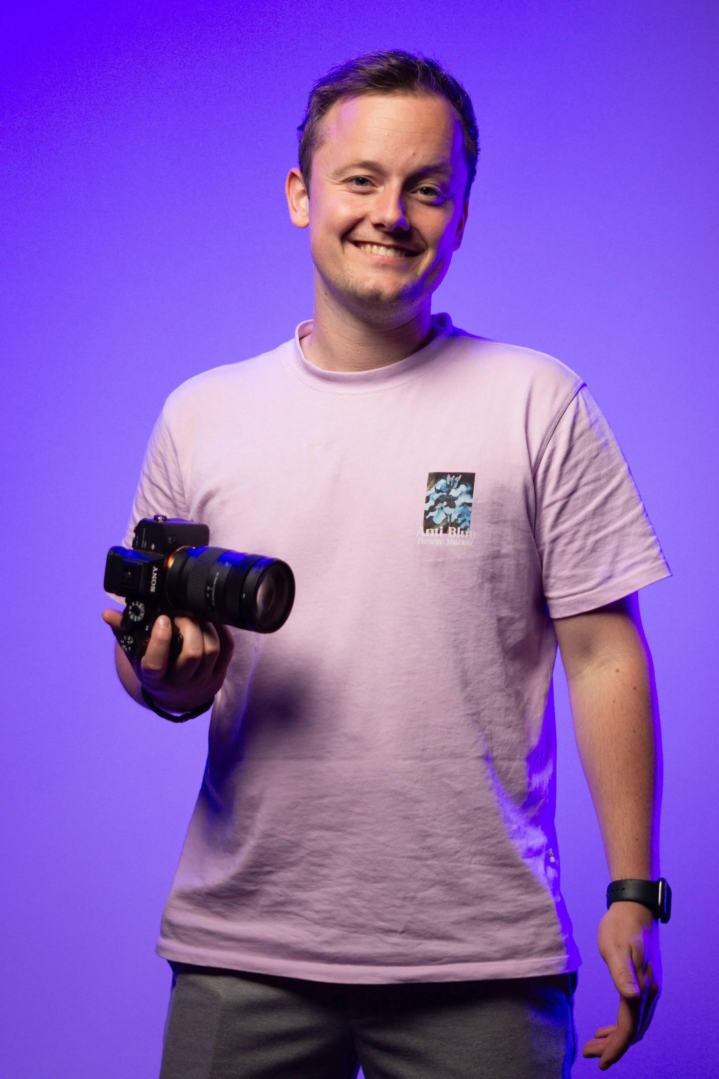 Man die een professionele camera vasthoudt, draagt een lichtroze T-shirt met een print van bloemen en tekst, staande voor een paarse achtergrond.