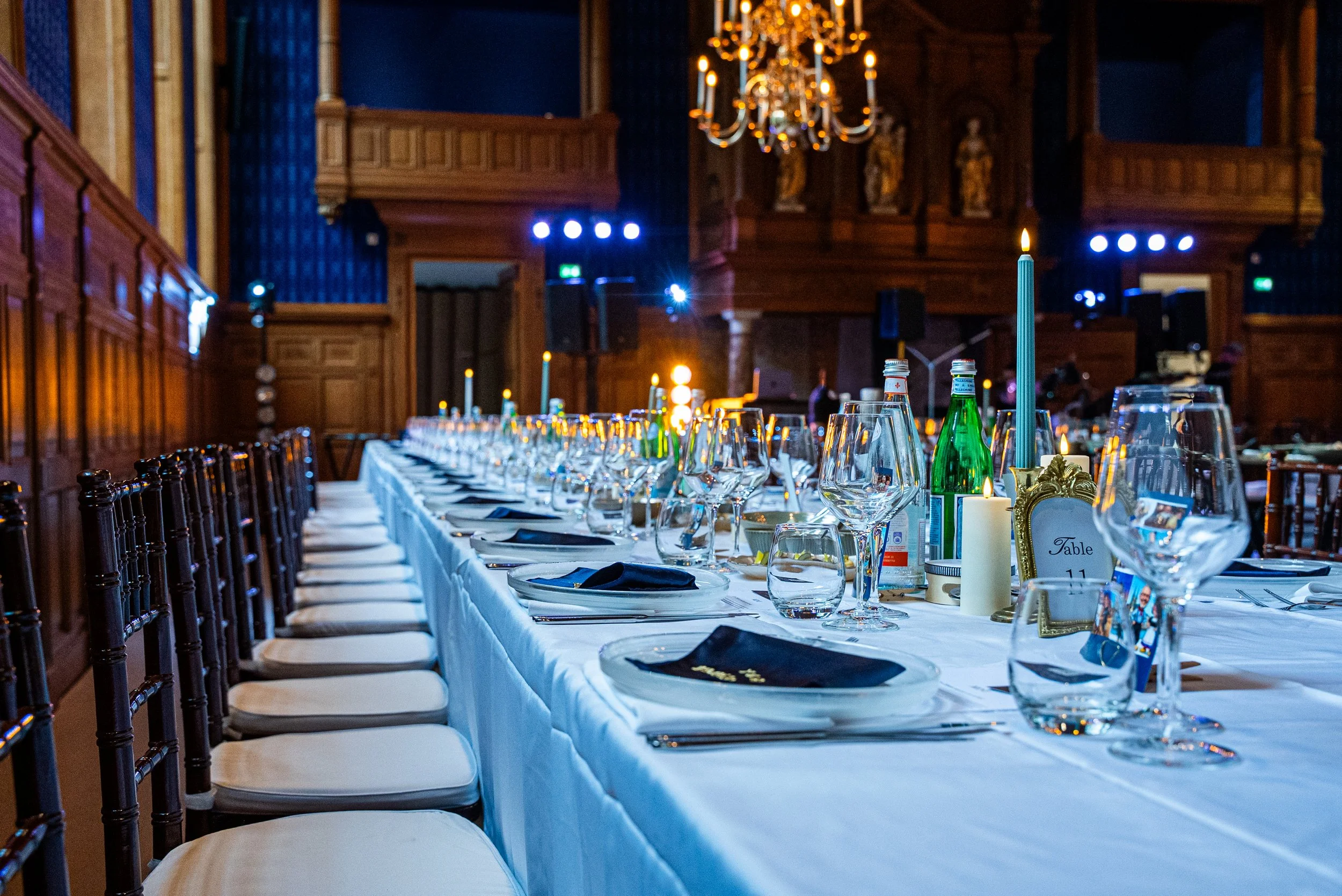 Een lange feesttafel versierd met glazen, borden, bestek en kaarsen, in een grote, elegante kamer met houten wandbekleding en een grote houten kroonluchter aan het plafond.