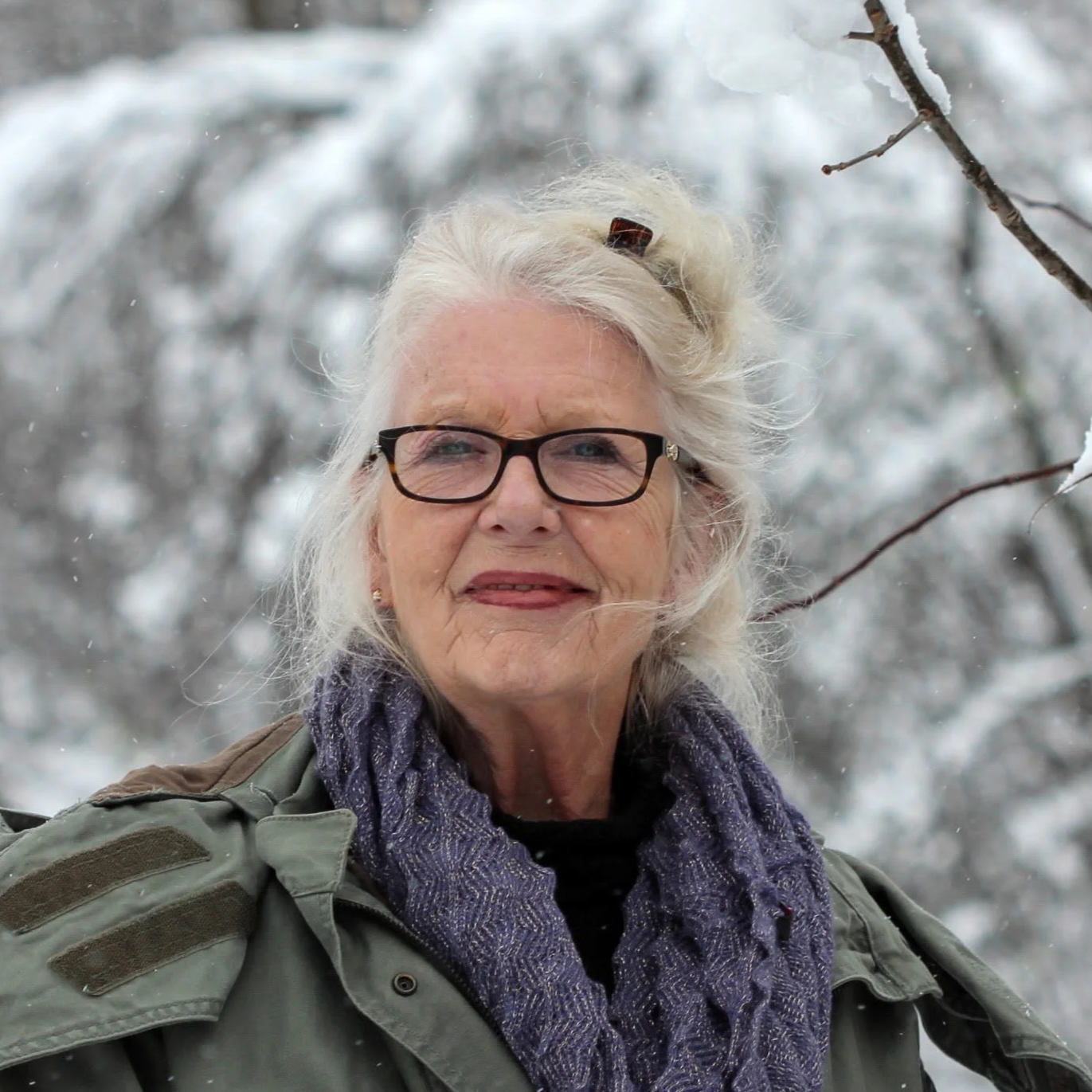 A smiling elderly woman with glasses and grey hair standing outdoors in a snowy landscape, wearing a purple scarf and a green jacket.