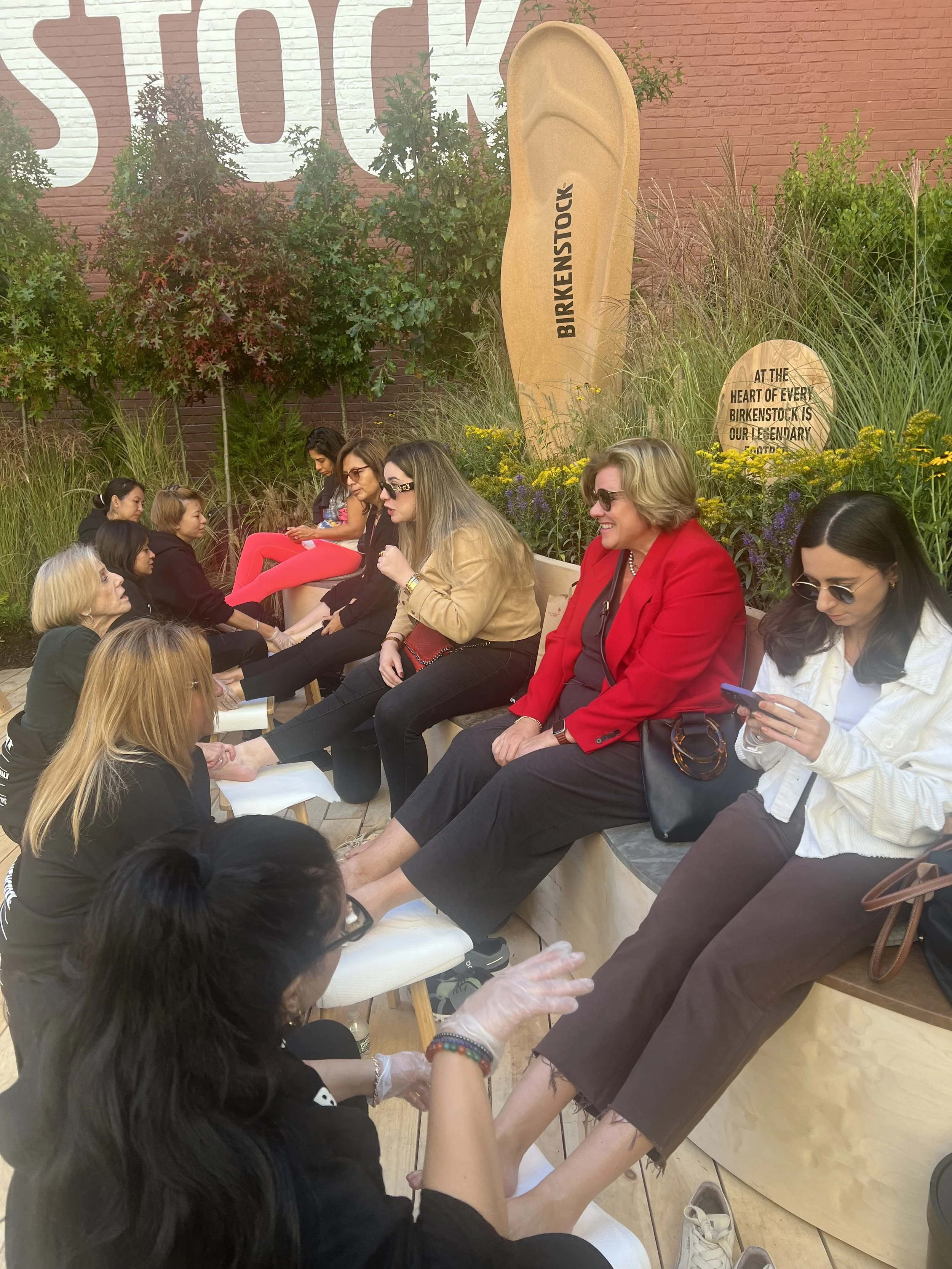 Women sitting in a row at a foot spa, receiving pedicures, outdoors with greenery and a large Birkenstock sign in the background.