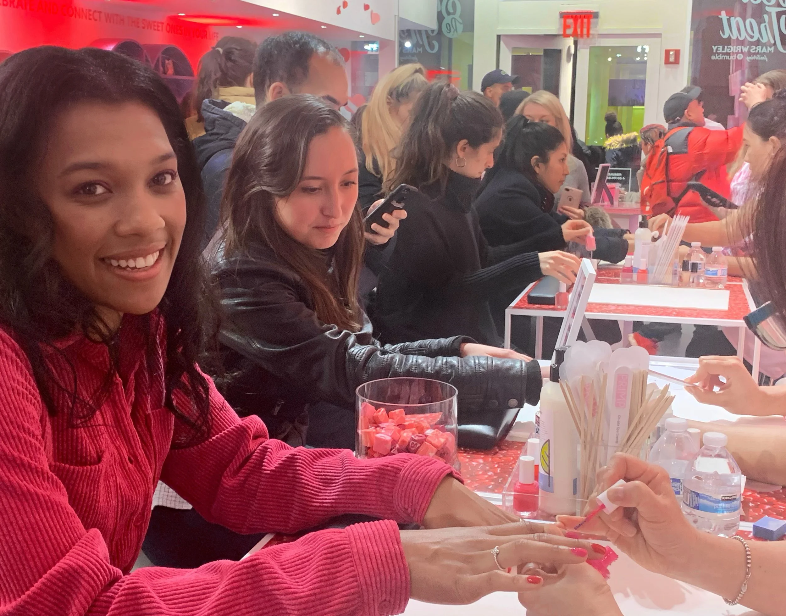 Young woman smiling with dark curly hair, wearing a red sweater, receiving a manicure at a nail salon surrounded by other clients getting services, with manicure tools and drinks on the table.