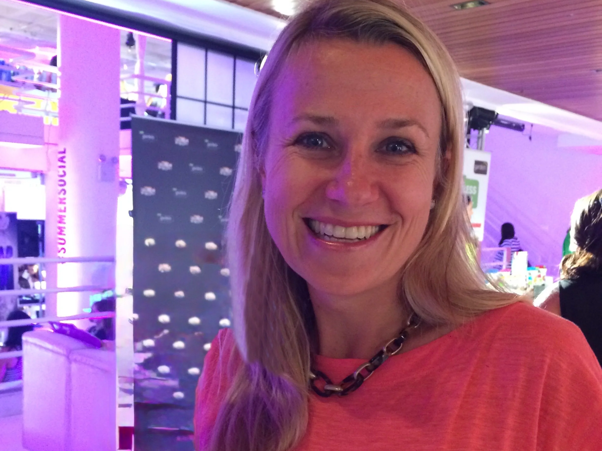 A smiling woman with blonde hair wearing a red top and a black chain necklace at an indoor event with purple lighting and a black backdrop.
