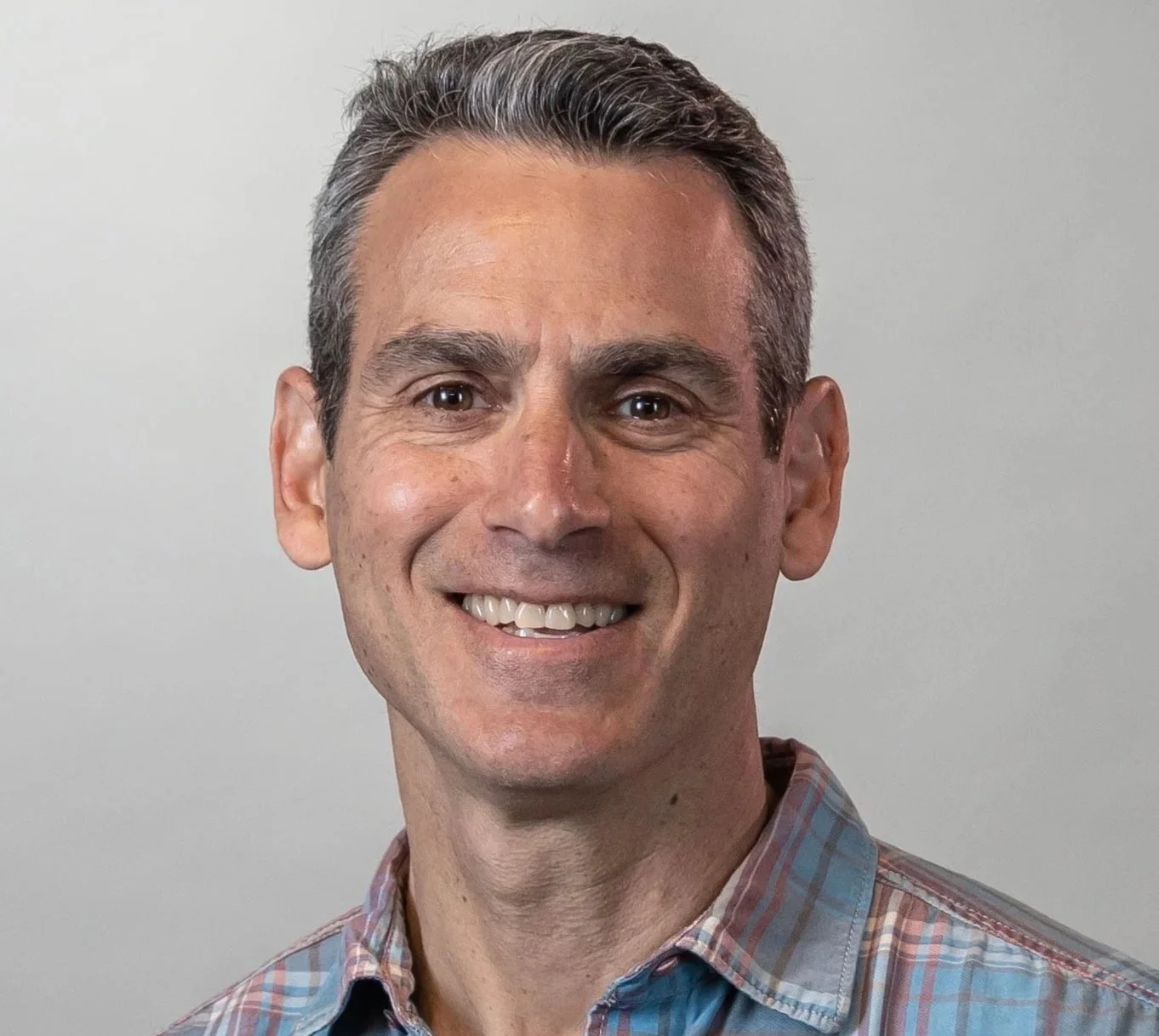 A smiling man with short, dark hair and light skin, wearing a plaid shirt, standing against a plain, light-colored background.