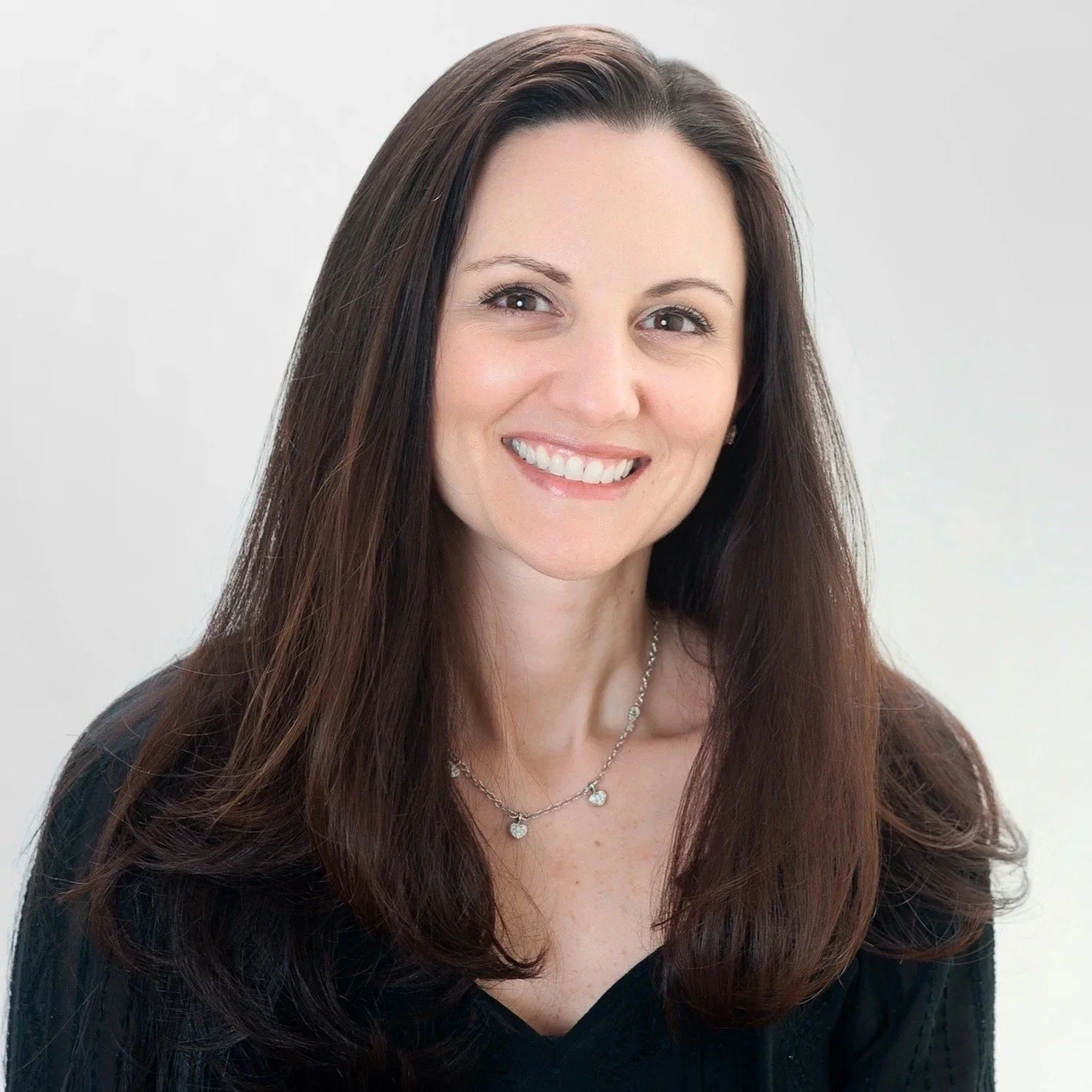 Professional portrait of a woman with long brown hair, smiling, wearing a black top and a delicate silver necklace with small charms, against a light gray background.