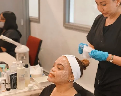 A woman receiving a facial treatment at a spa or beauty clinic, with a skincare mask applied and a headband, while a professional applies the product with gloves nearby.