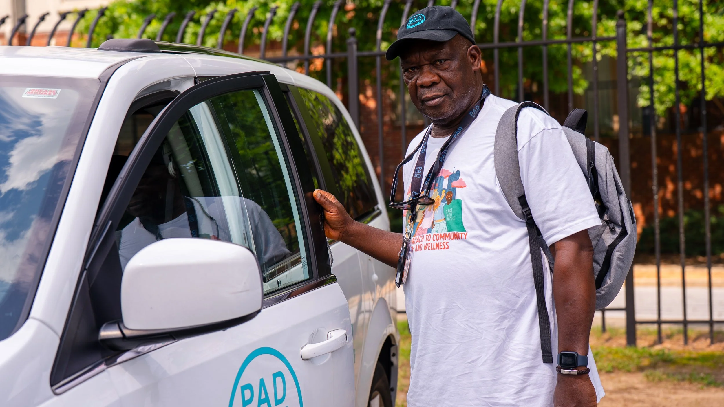 Kasali Obanoyen, Community Responder Team Lead stands next to a white PAD van, with an open door. He wears a white PAD T-shirt and a gray backpack.