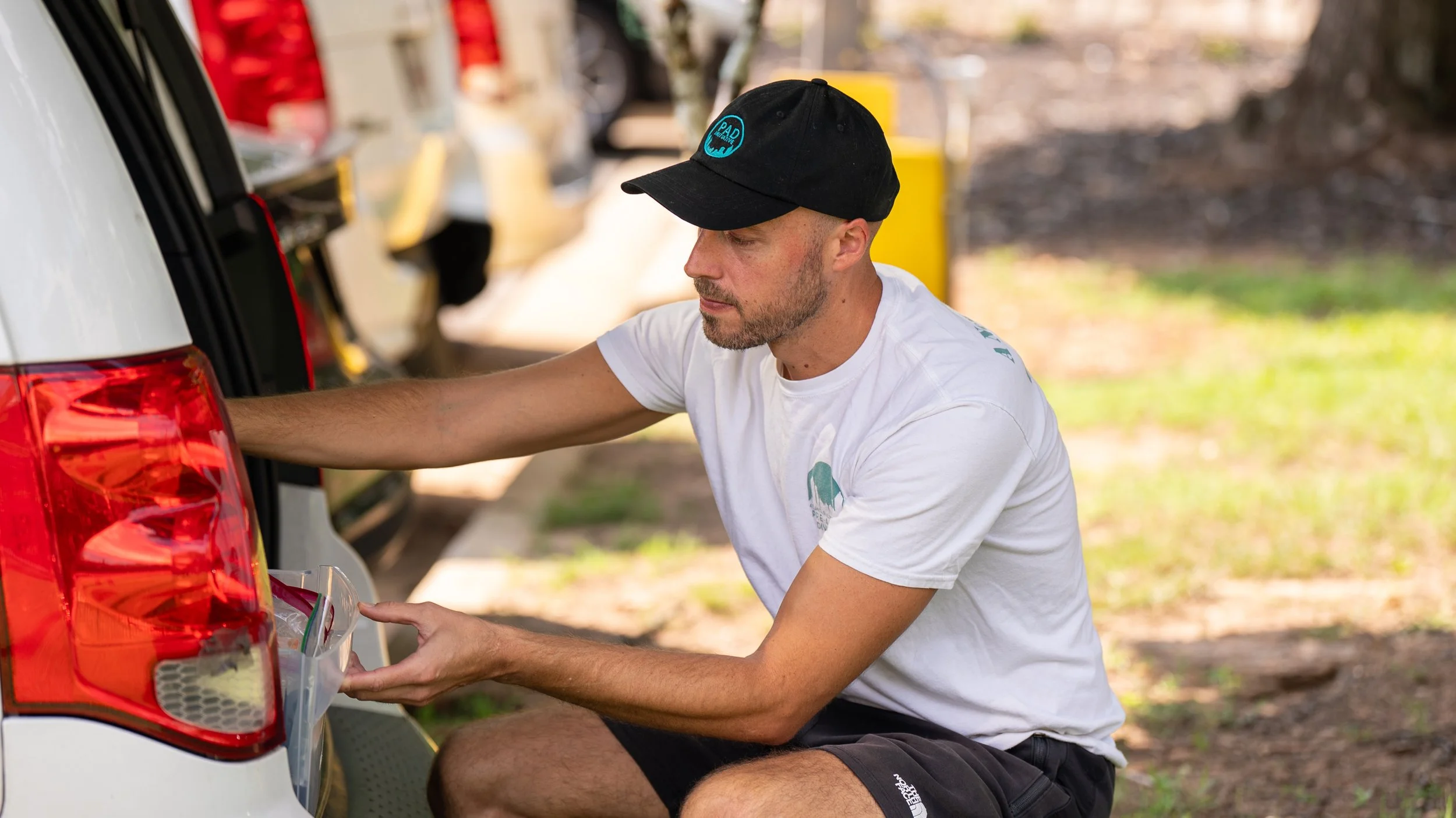 Image features Thaddeus Pettrey, a PAD community responder, wearing a white t-shirt and black PAD hat, squatting behind a PAD van, placing an item into a box in the van's trunk.