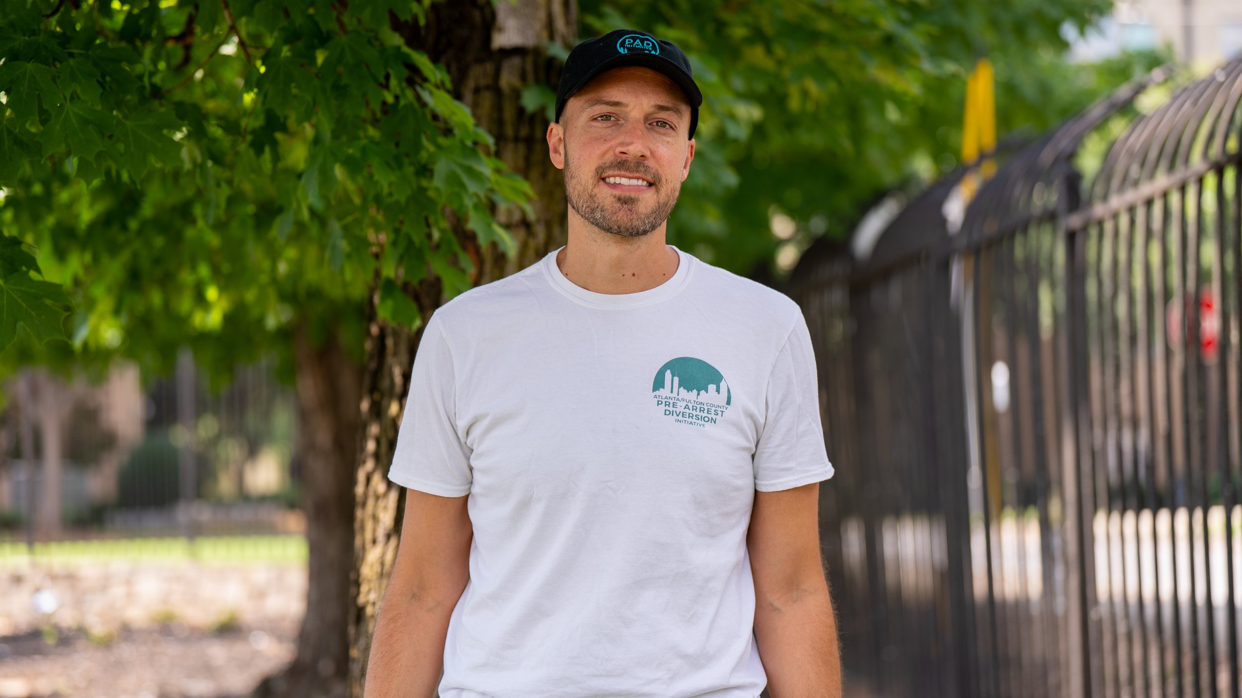 Headhsot of Thaddeus Pettrey, a community Responder at PAD. Thaddeus wears a black PAD hat and a white t-shirt with teal text and graphics that reads:"ATLANTA/FULTON COUNTY PRE-ARREST DIVERSION INITIATIVE".Thaddeus stands in front of a tree.